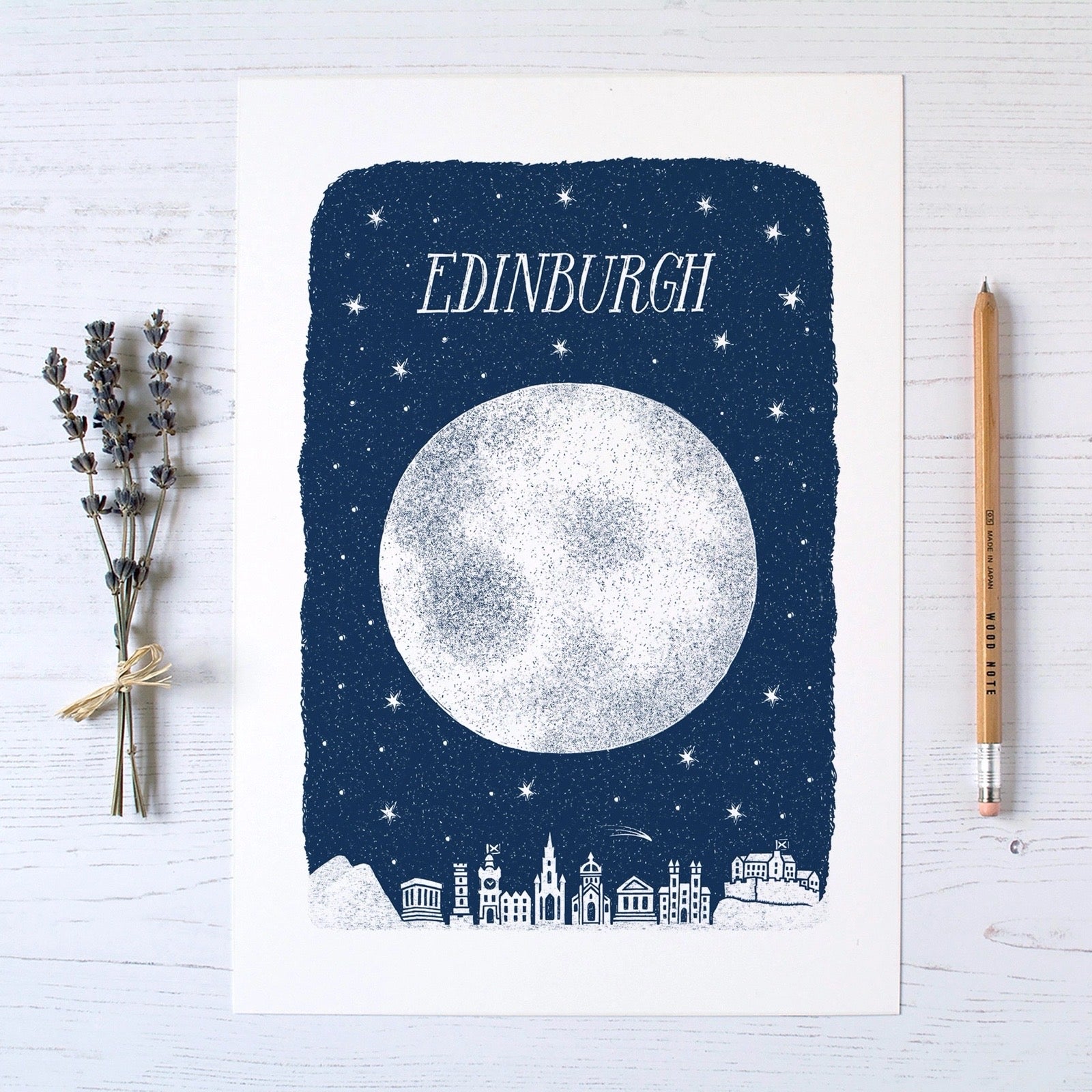 Poster with moon and cityscape design on a blue background, featuring the word 'Edinburgh', on a table next to a pencil and lavender sprig.