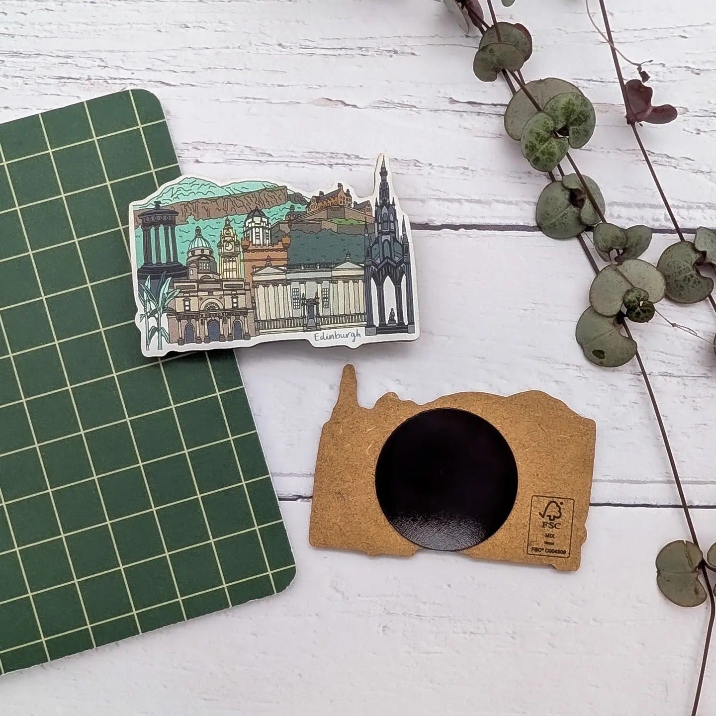 A laser-cut wooden magnet featuring a line drawing of Edinburgh's iconic buildings, including Edinburgh Castle and The Scott Monument, with another magnet turned over, on a white and green background with a plant.