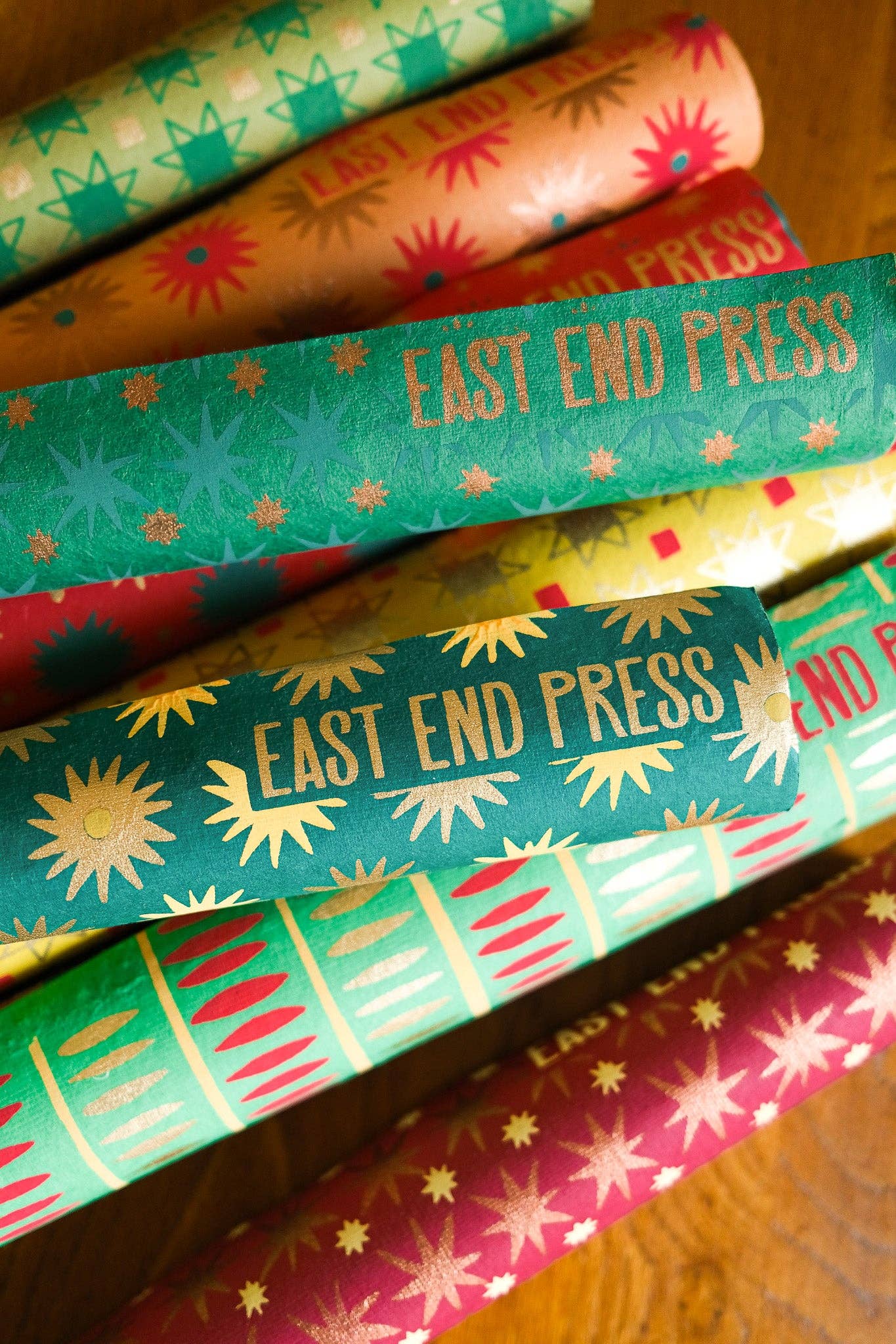 A close-up of several rolled sheets of patterned wrapping paper stacked on a wooden surface. The paper features retro starburst and geometric designs in green, orange, yellow, red, and pink, with 'EAST END PRESS' printed in gold foil on multiple rolls.
