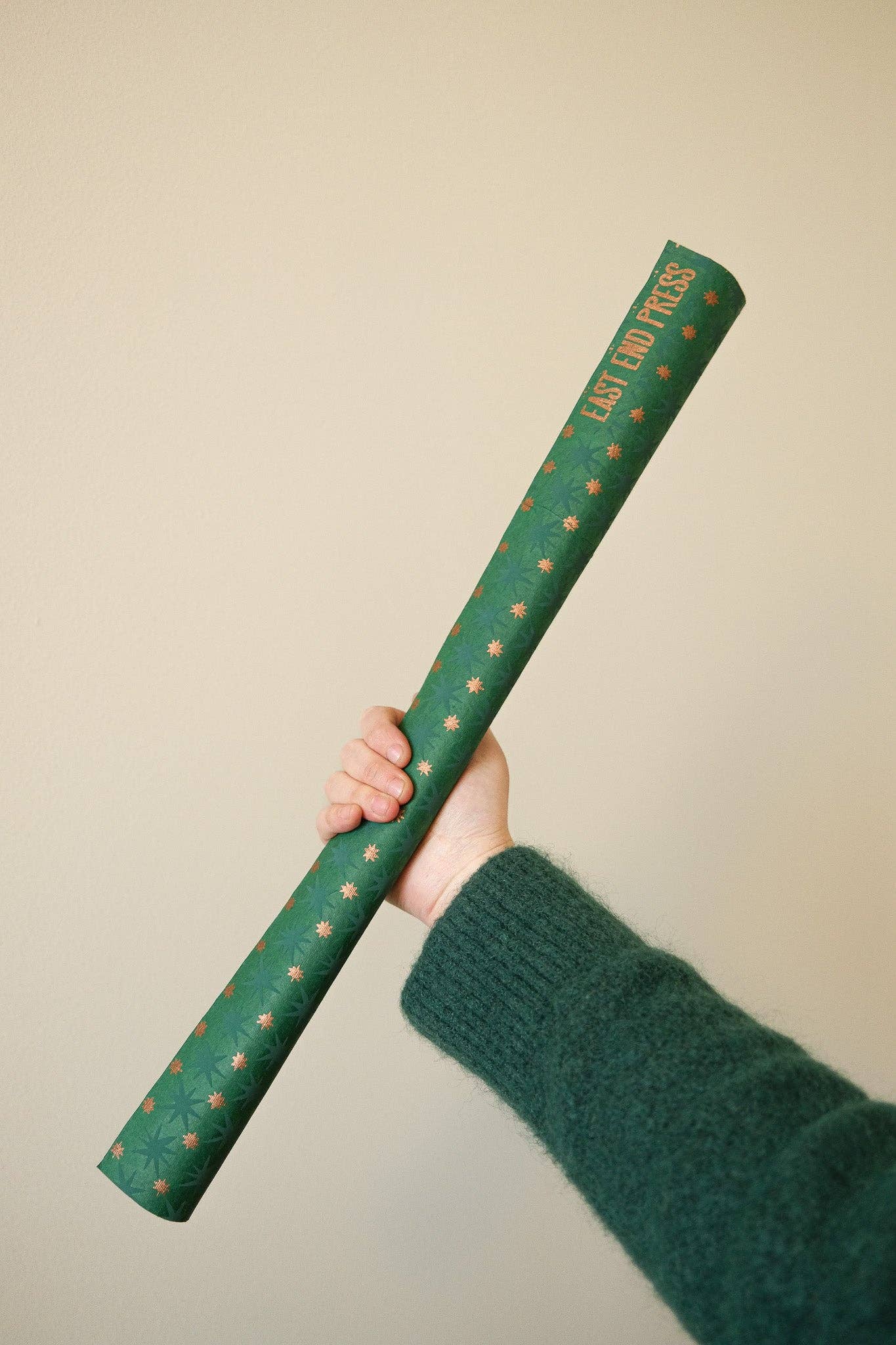 A hand holding a rolled-up green wrapping paper tube with small gold stars printed on it, against a plain beige background.