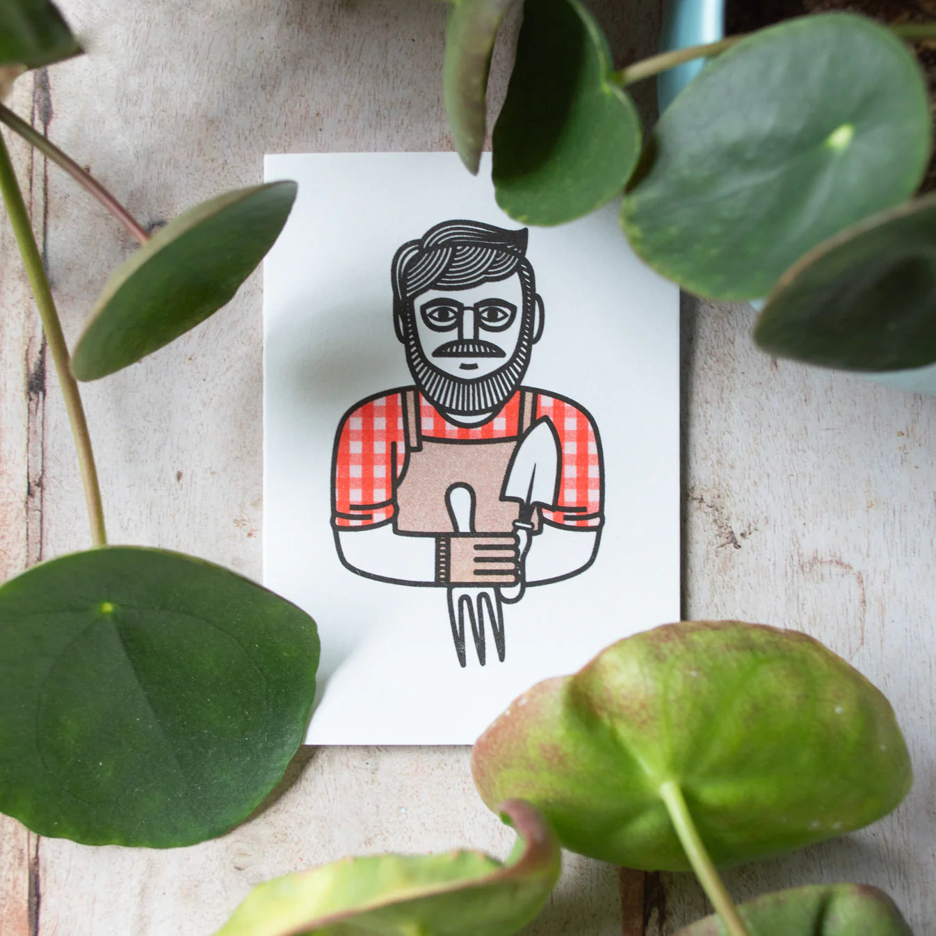 Illustration of a gardener on a white card surrounded by green leaves on a wooden surface.