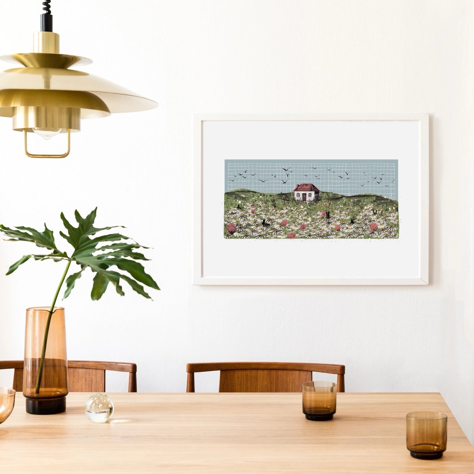 Dining room with a framed artwork of a house on a hill, wooden table, chairs, and decorative items.