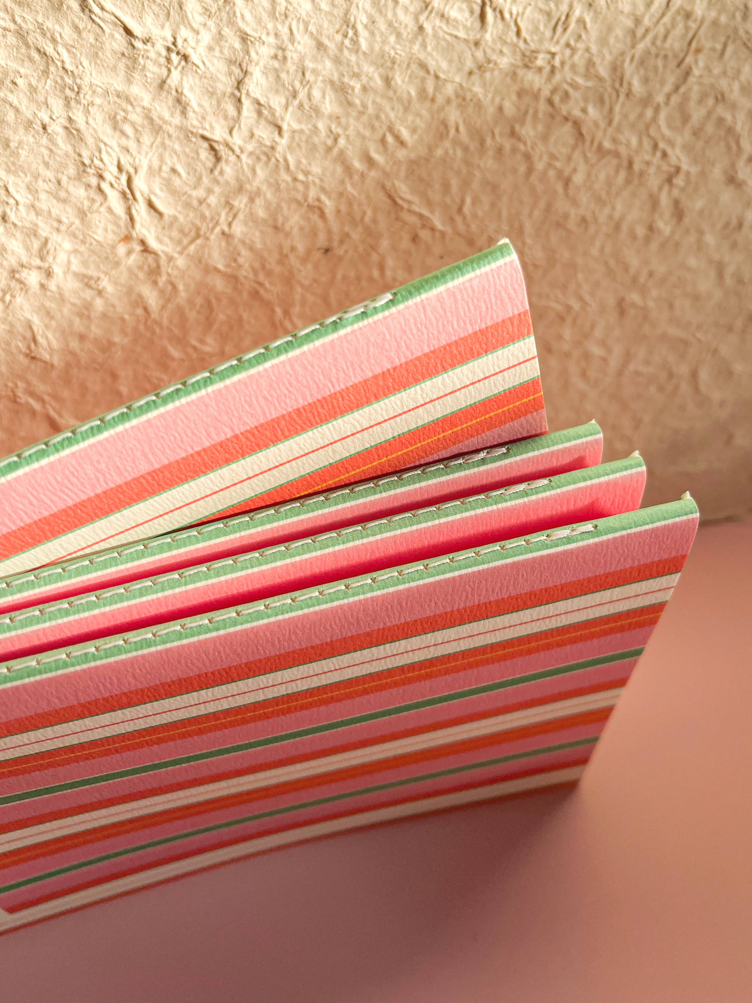 A stack of striped paper notebooks with pink, orange, green, and white horizontal bands, standing on a dusty pink surface against a textured beige background.