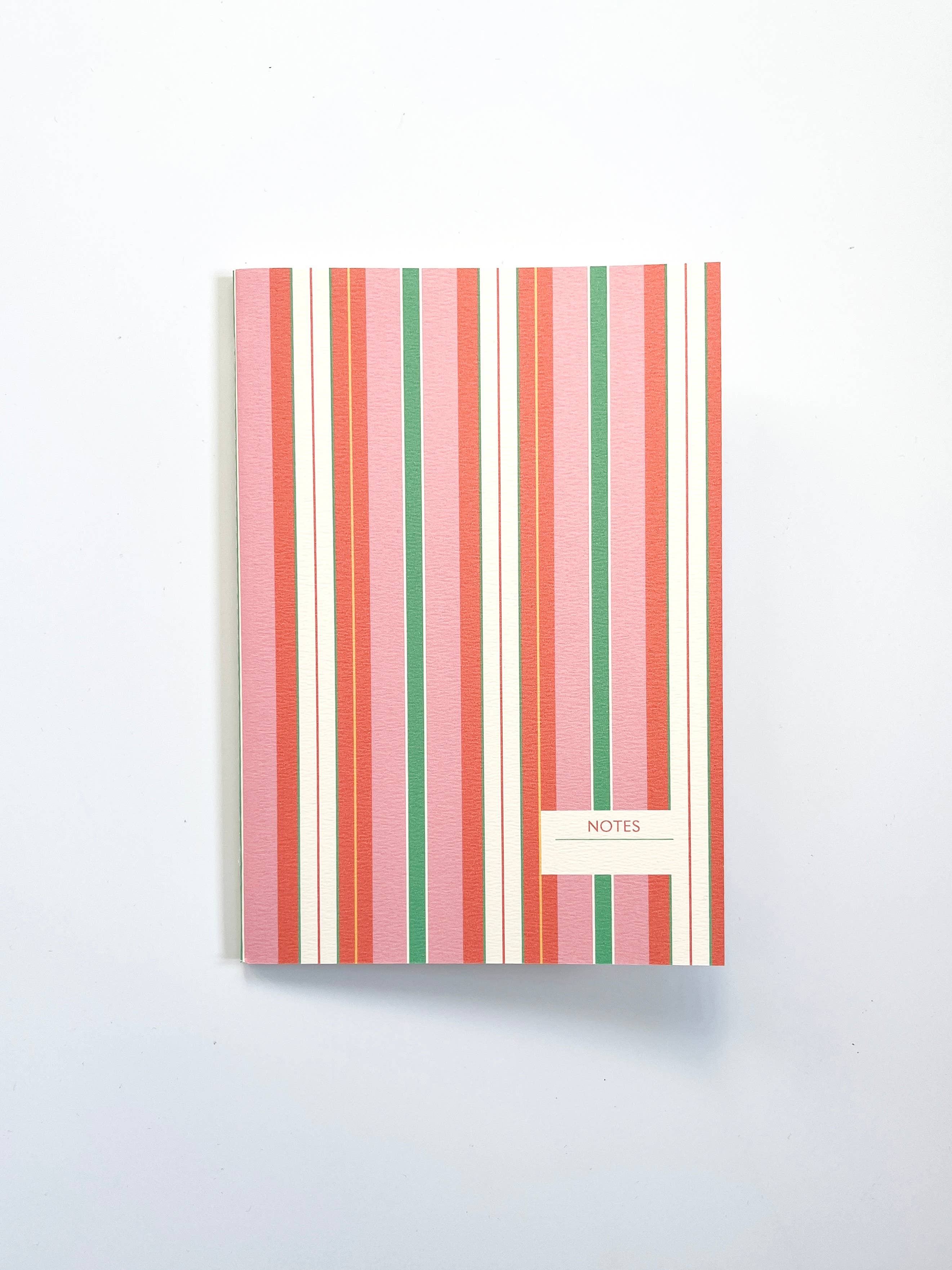 A notebook with a vertical striped cover in shades of pink, coral, green, and cream, featuring a small cream label at the bottom right that reads 'NOTES'.