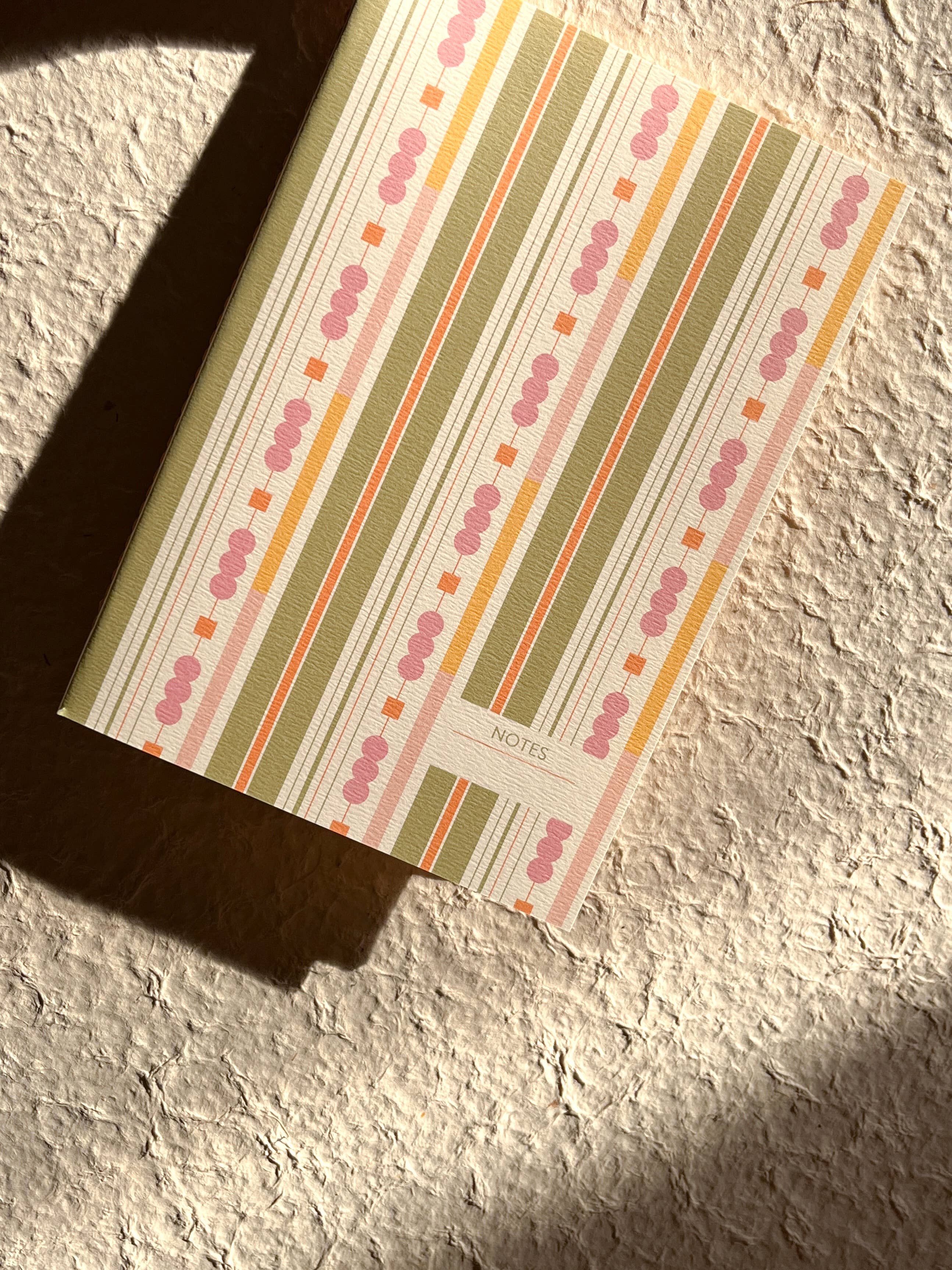 A notebook with a retro striped cover pattern in pink, orange, olive green, and cream, resting on a textured beige surface with a strong diagonal shadow.