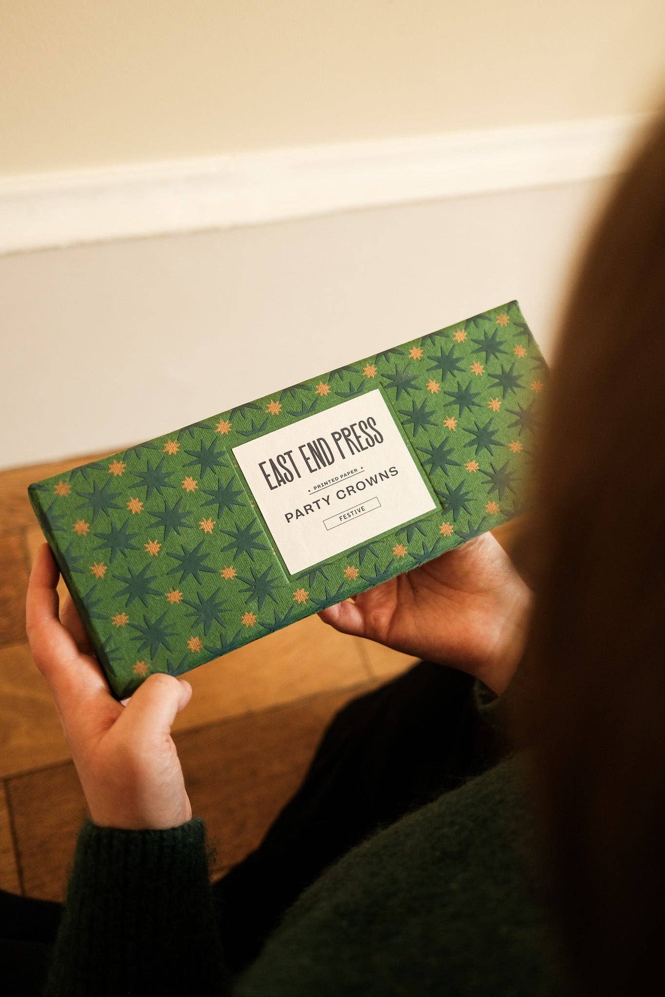 A person holds a green patterned box labeled 'East End Press London Party Crown' in an indoor setting.