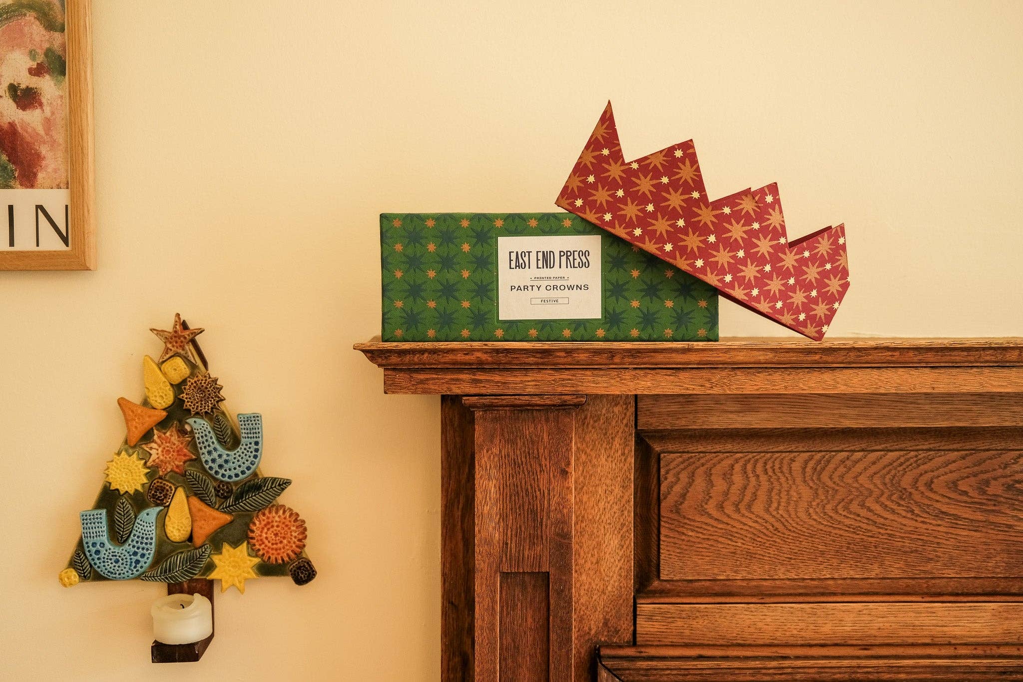 A green patterned box with a white label reading 'EAST END PRESS' sits on a wooden mantel, with a red patterned paper crown leaning against it. The scene is indoors against a warm cream wall, with part of a framed print and a decorative wall tree visible.