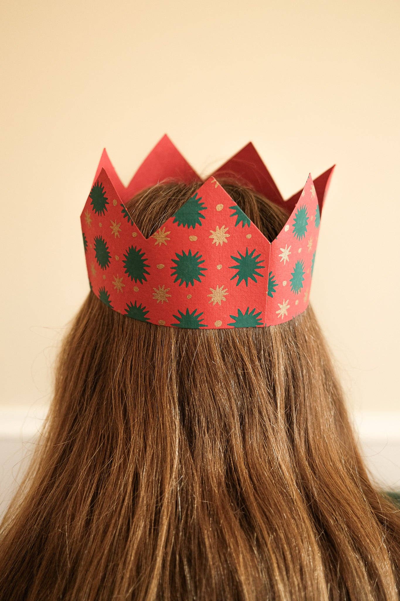 A red paper crown with green and gold starburst patterns worn on the head of a person with long brown hair, photographed from behind against a plain beige wall.