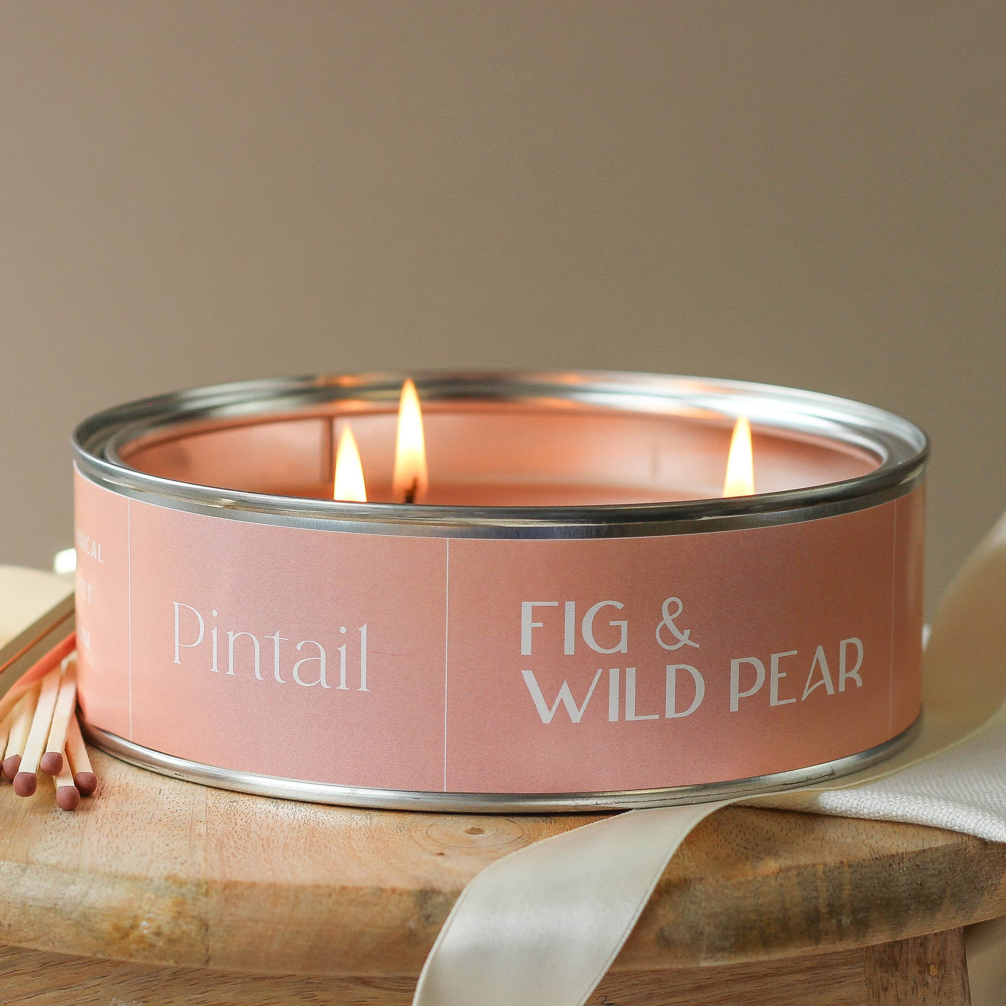Candle in a pink tin labeled 'Pintail Fig & Wild Pear' on a wooden surface.