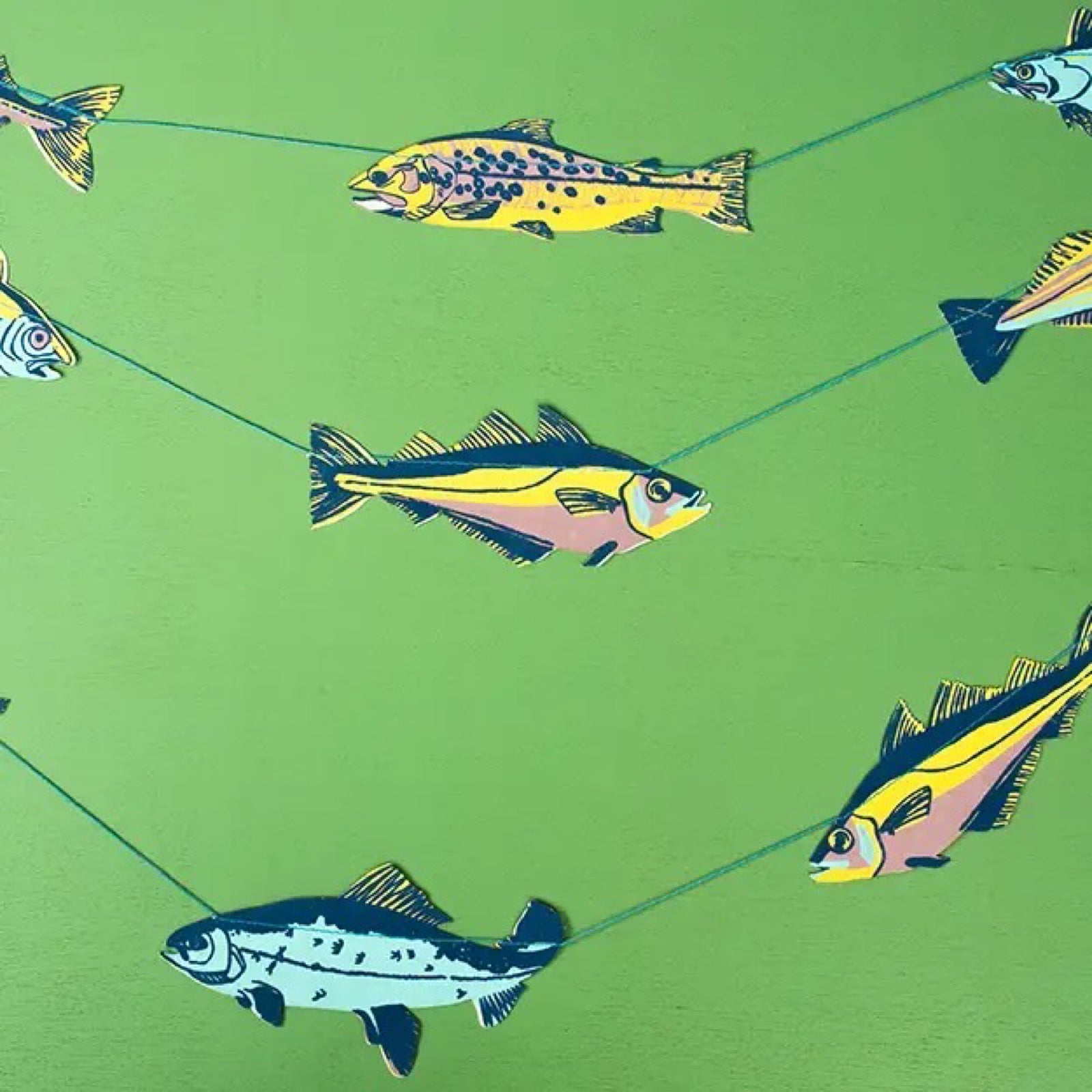 Fish paper garland with 10 screen-printed fish in shades of pink, turquoise, navy and yellow hanging on a green wall.
