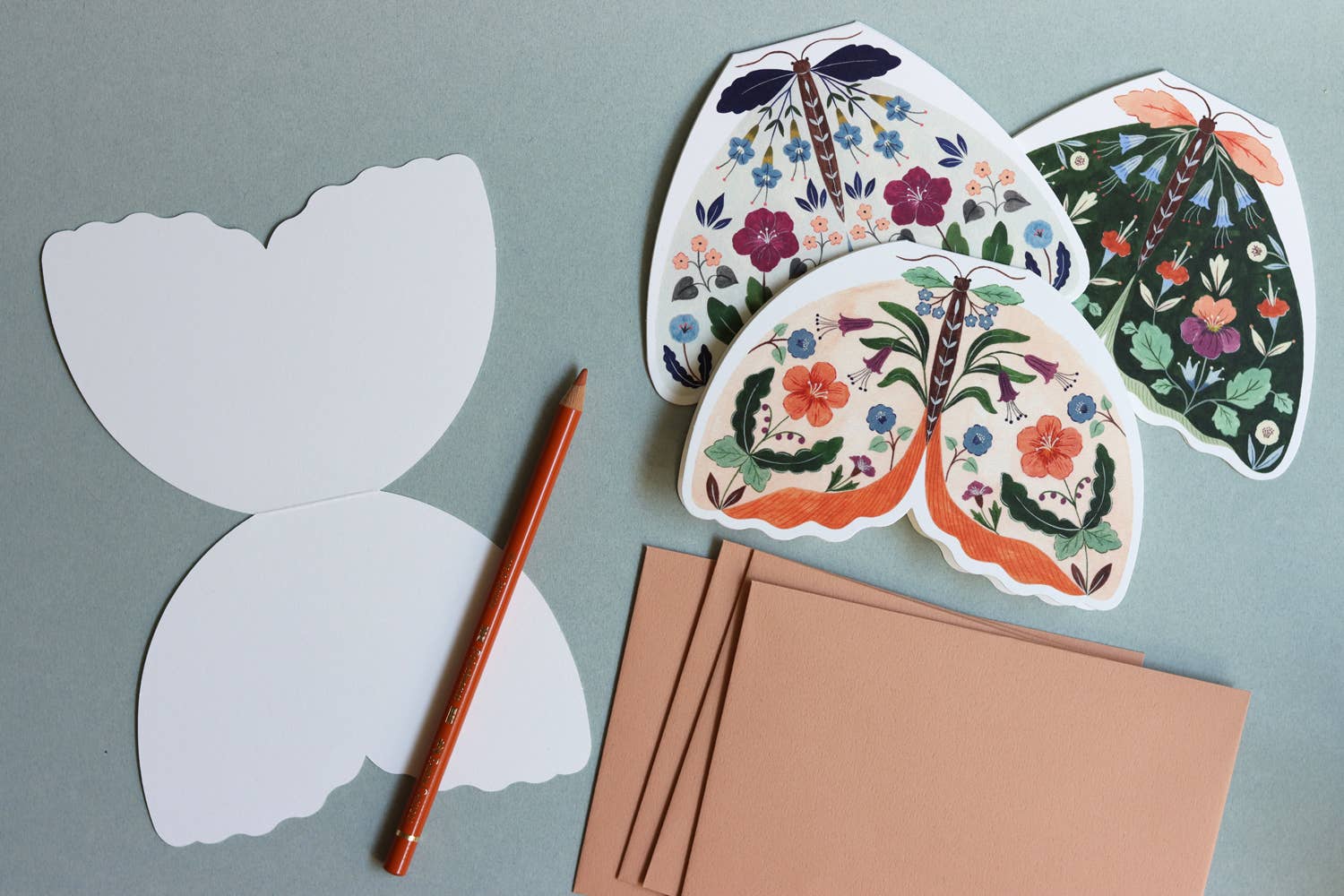 Moth card paper cutouts with floral patterns, a pencil, and brown paper on a grey background.
