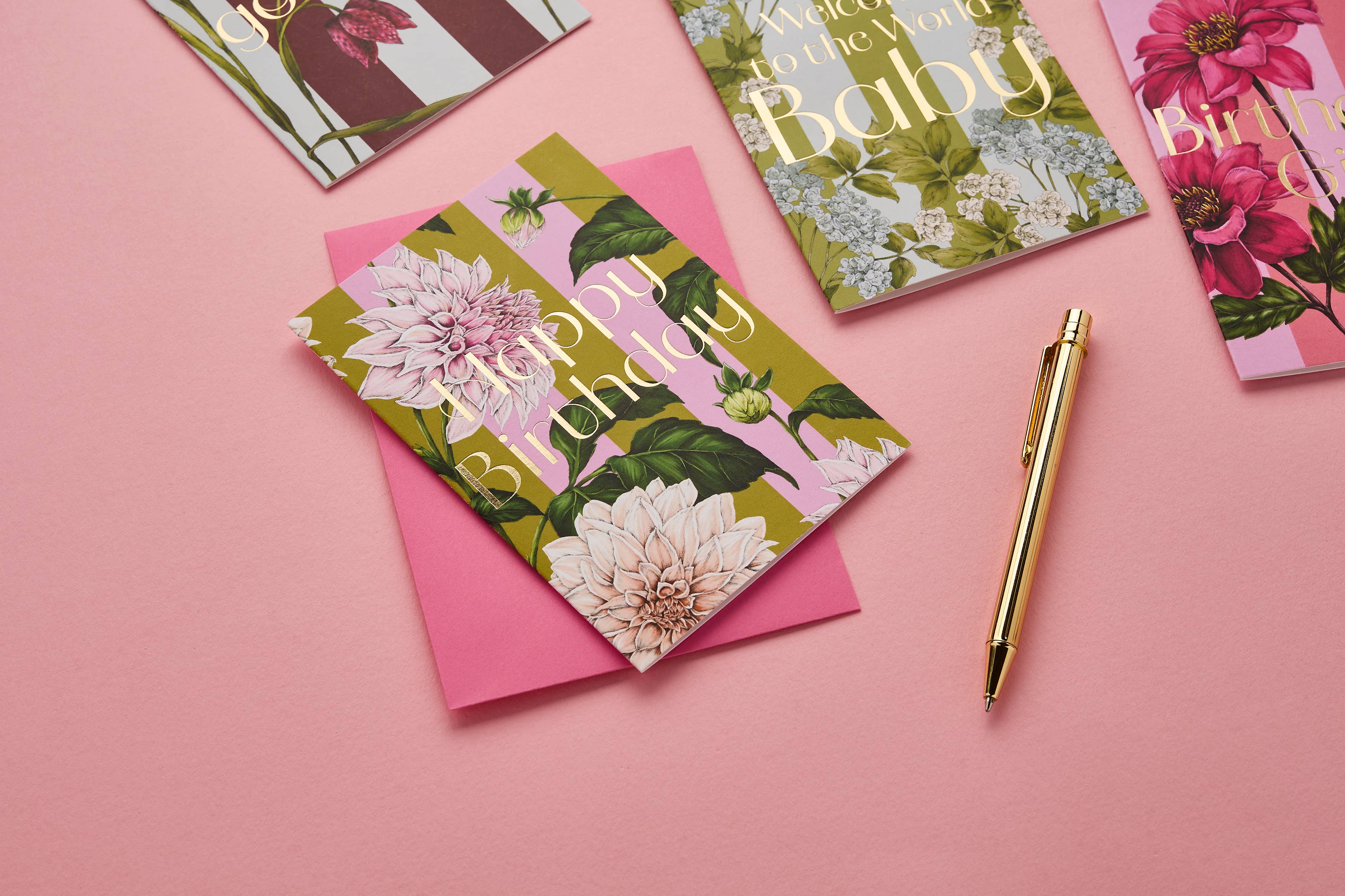 A styled flat-lay of floral greeting cards and envelopes on a pink background, with a gold pen nearby. The main visible product is a greeting card with a green, pink, and white dahlia pattern and the words 'Happy Birthday' in gold script.