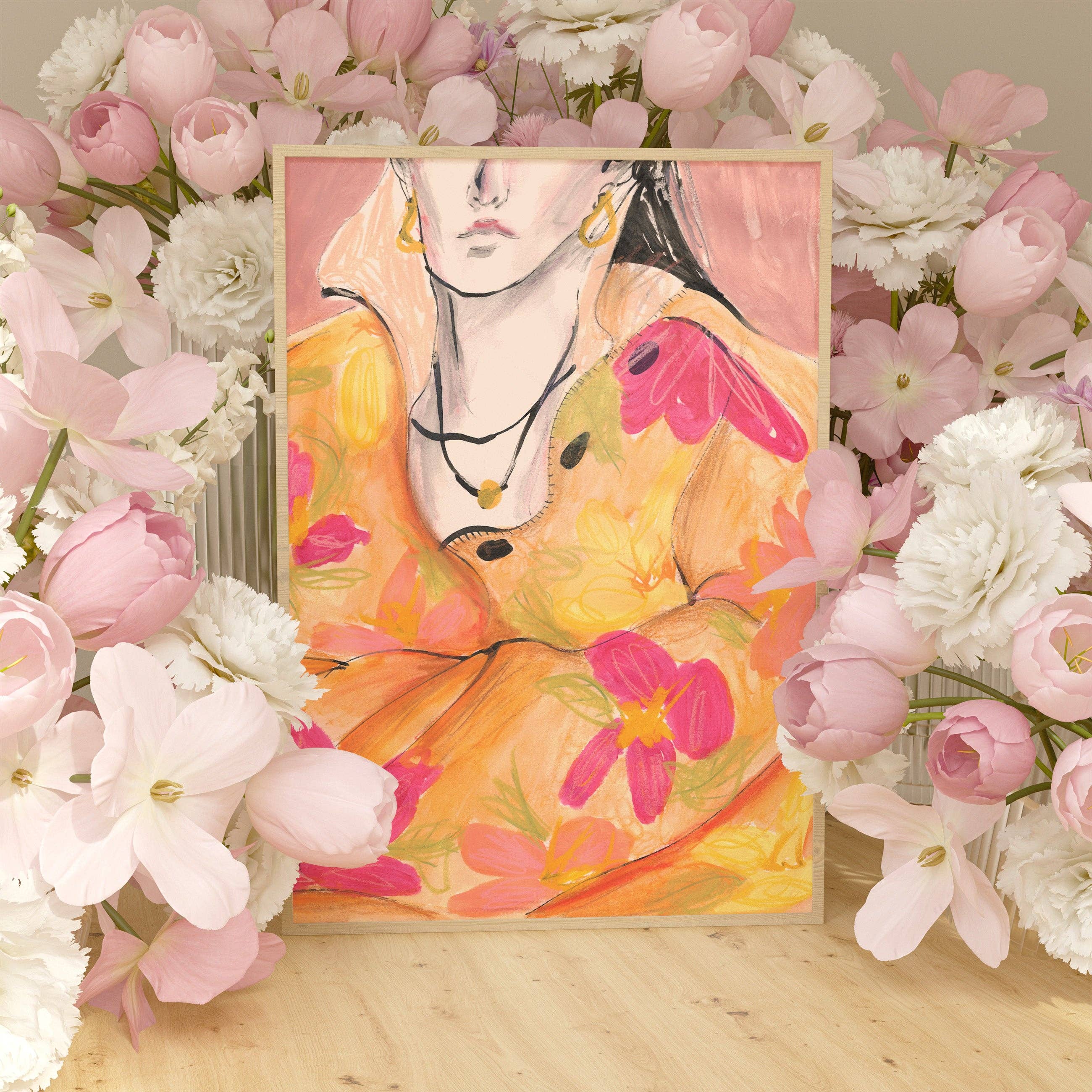 A framed art print featuring a stylised portrait of a woman in a yellow dress with pink floral accents, displayed on a wooden surface and surrounded by pink and white flowers.