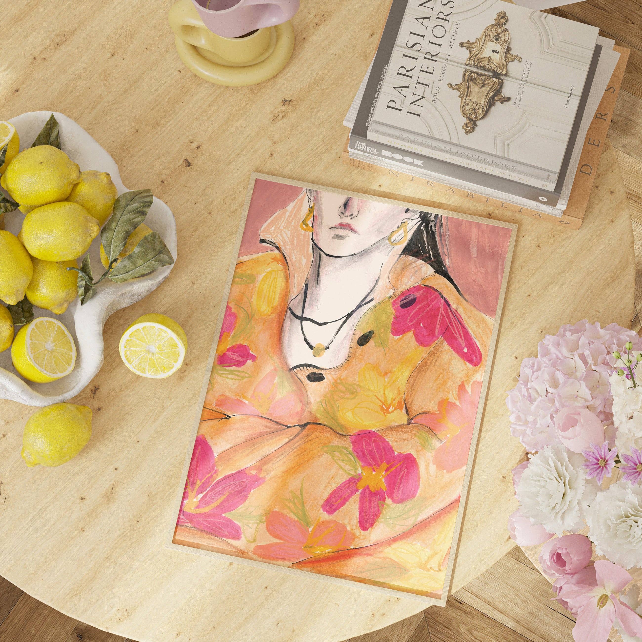 A framed colorful painting of a woman in a floral dress lies on a light wood table, surrounded by lemons, flowers, a magazine, and a cup. The painting features pink, yellow, orange, and black tones.