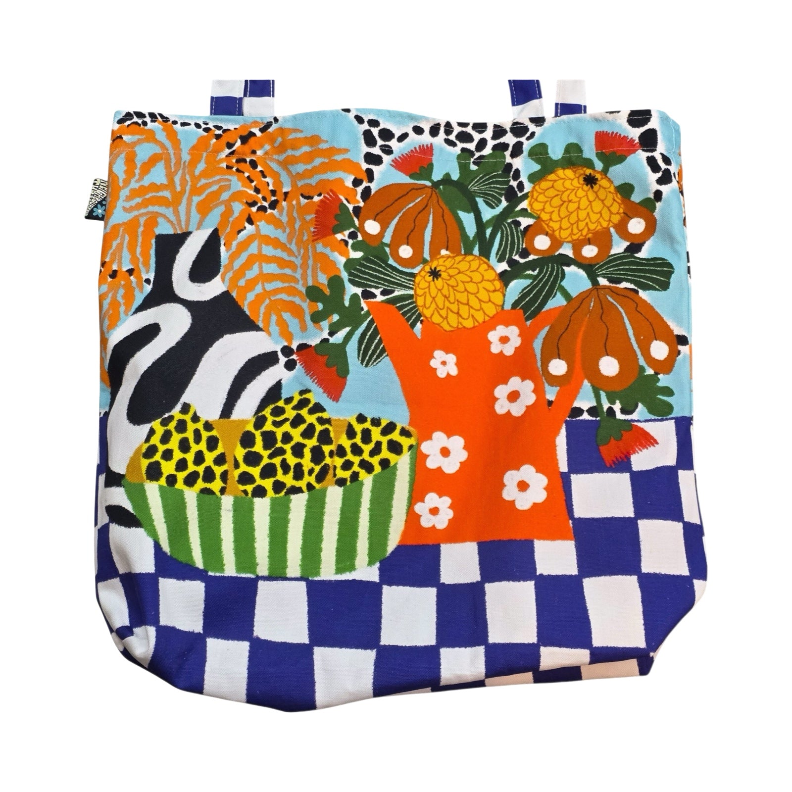 A bright and colourful tote bag featuring a black and white vase with orange leaves, a red jug with white daisies full of colourful flowers and a blue and white checked base.