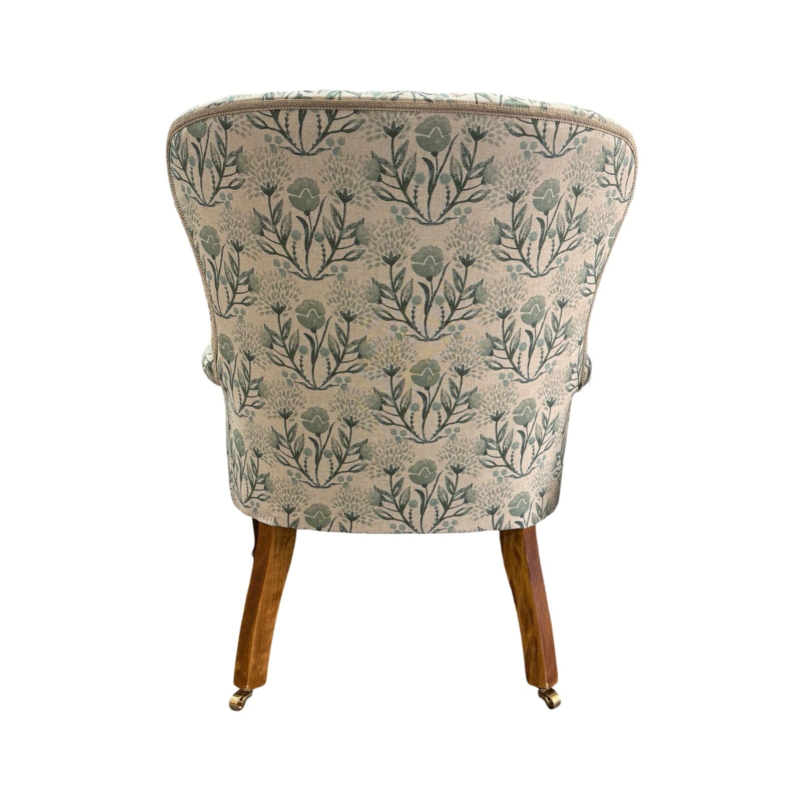 The back of an occasional armchair in cream linen fabric with flowers in soft blues and greens and finished with cream binding, mahogany stained legs and gold coloured castors.