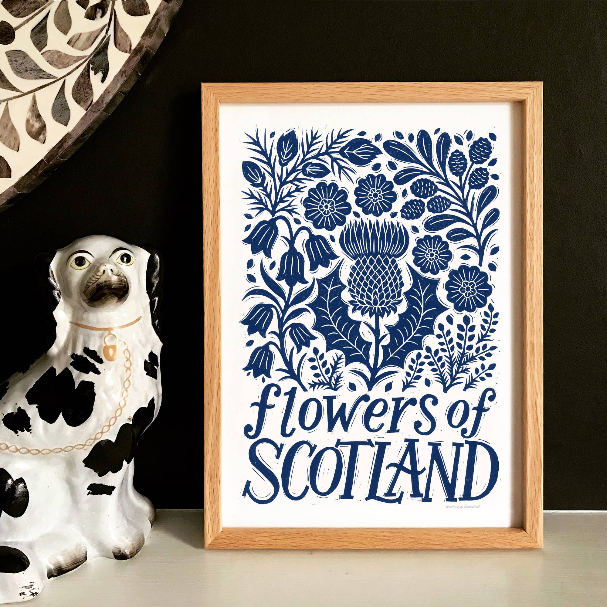 Framed artwork with blue floral design and 'Flowers of Scotland' text next to a ceramic dog figurine.