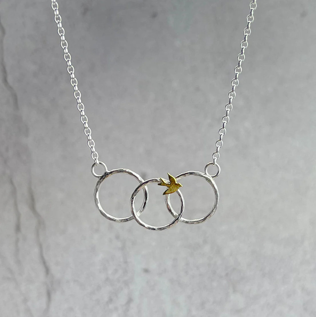 Silver necklace with three interlocking rings and a small yellow bird on a gray background.