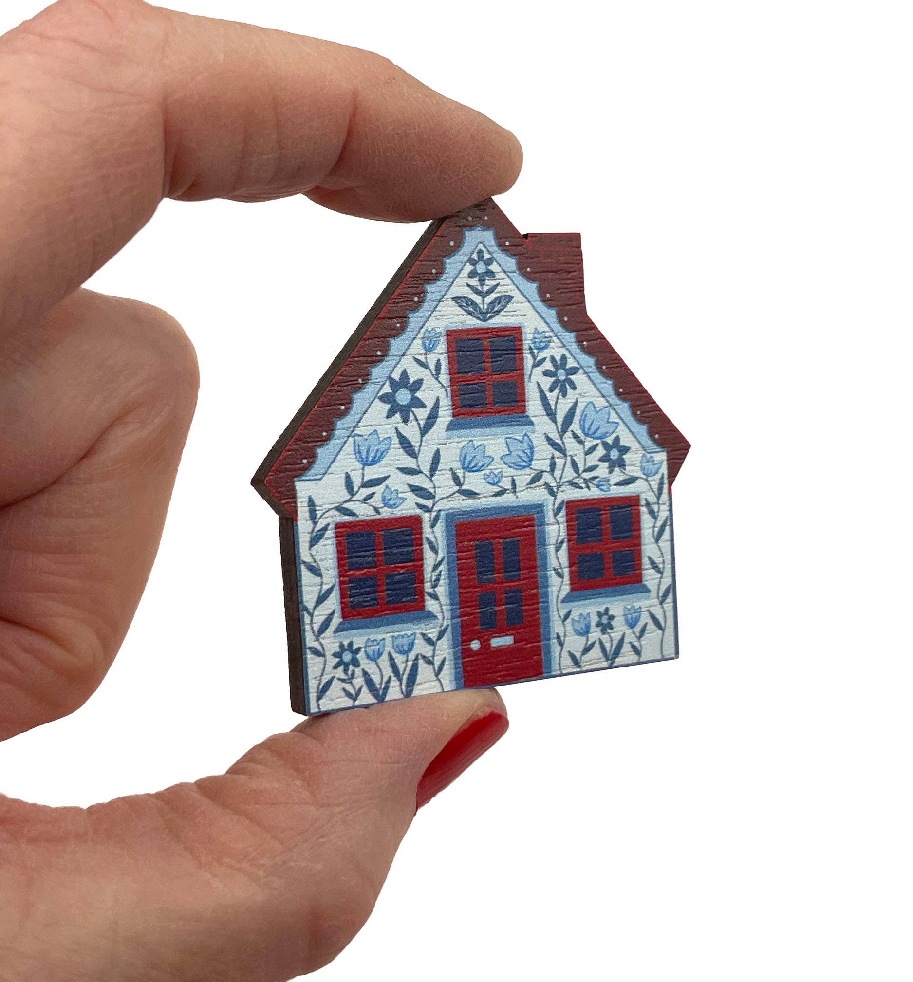 A hand holding a small decorative house-shaped item with a blue and white floral pattern, red door and window frames, and a brown roof against a plain white background.
