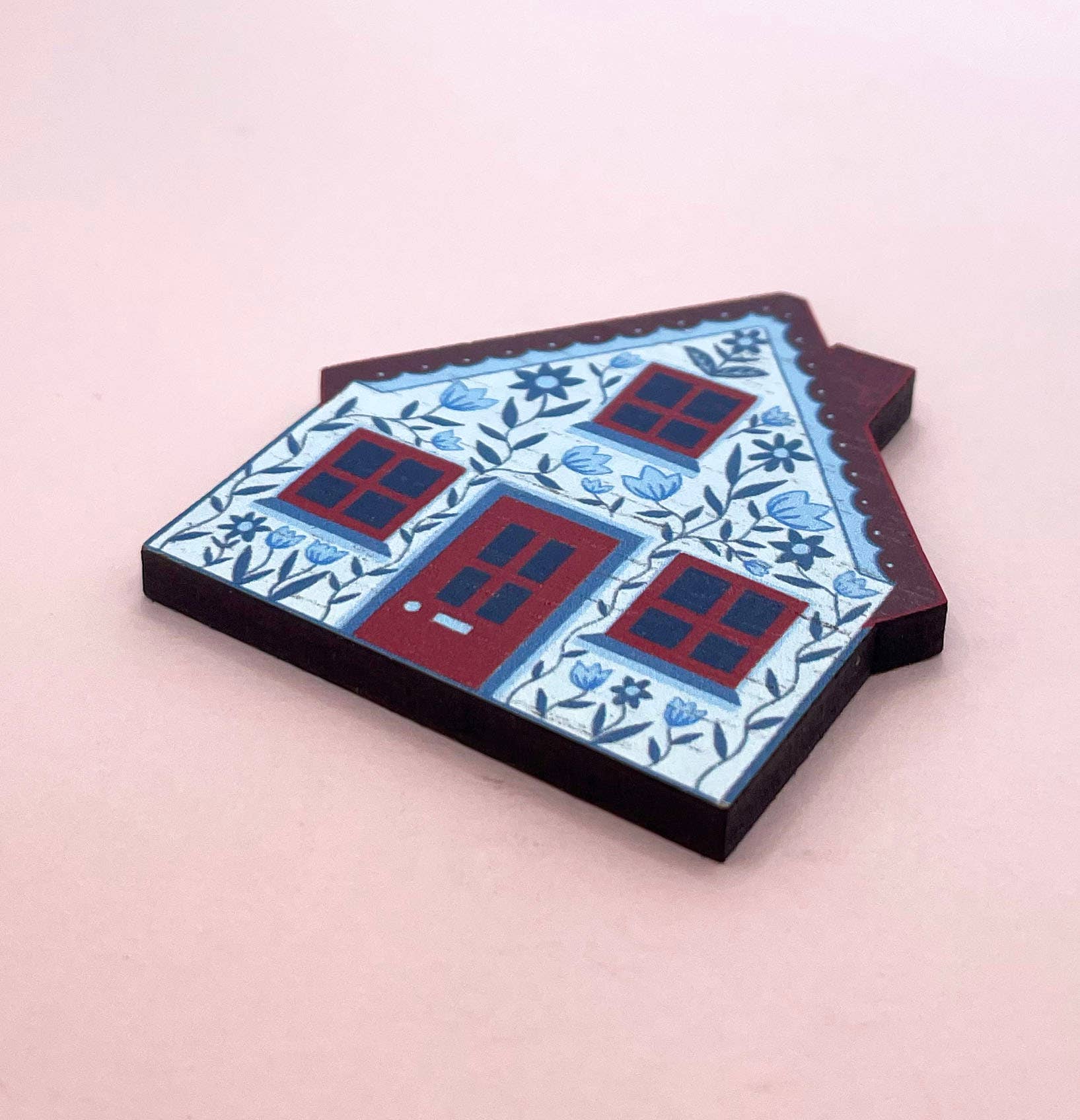 A house-shaped decorative item with a blue and white floral pattern and red window and door details, on a pink background.