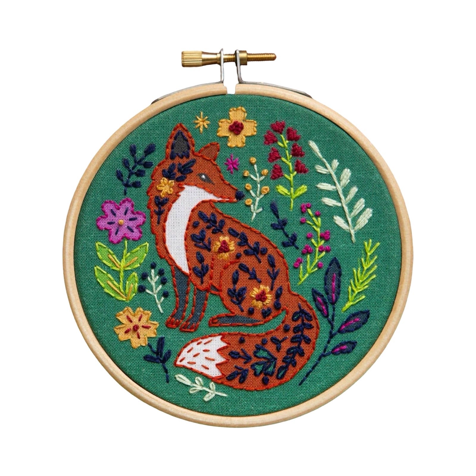 A circular wooden embroidery hoop holding a finished cross-stitch design of a fox surrounded by colourful flowers and leaves on a green fabric background, shown against a plain white background.