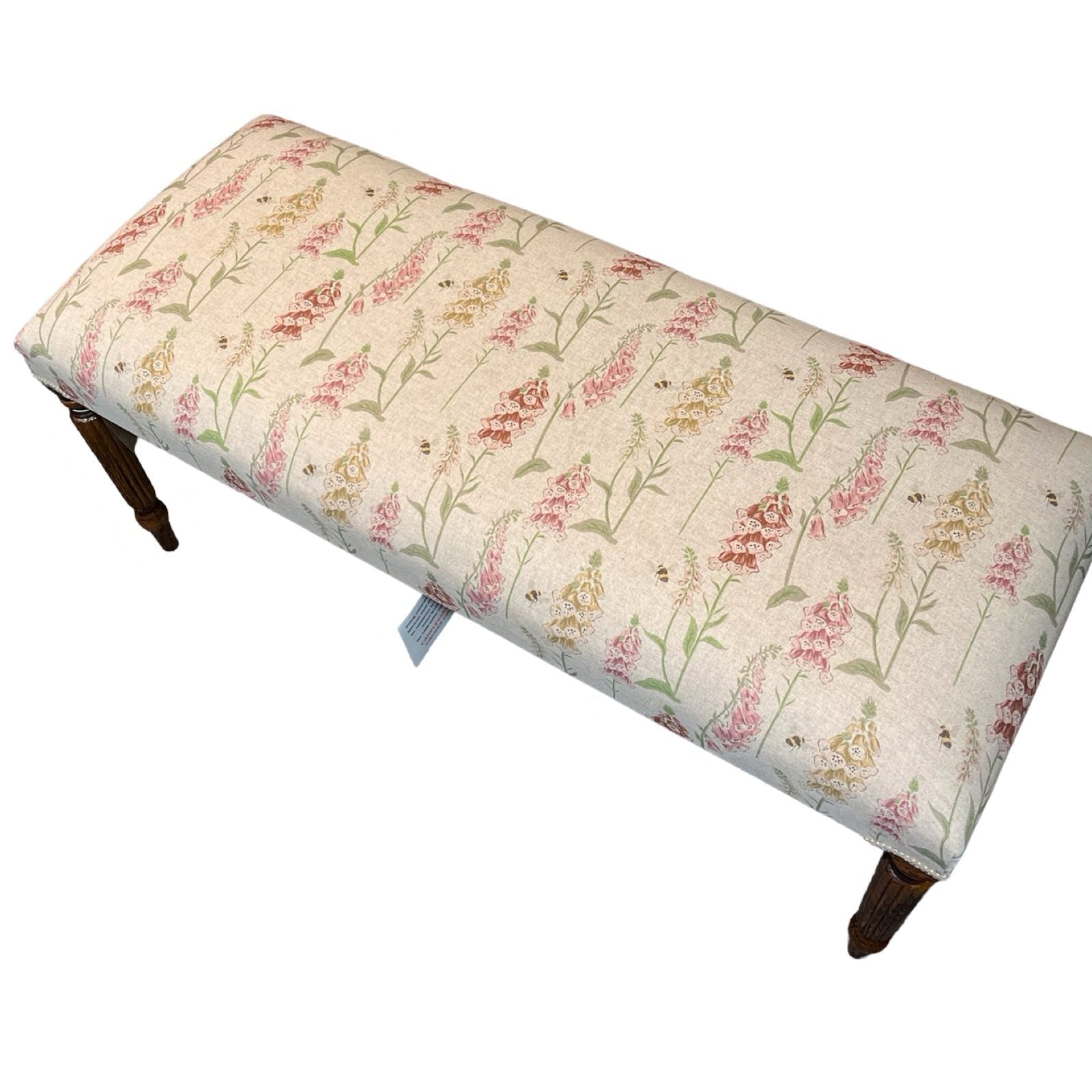 A rectangular bench with cream linen fabric upholstery featuring a floral pattern of foxgloves in pink, red, and yellow, with honey bees. It has mahogany legs and is finished with matching binding.