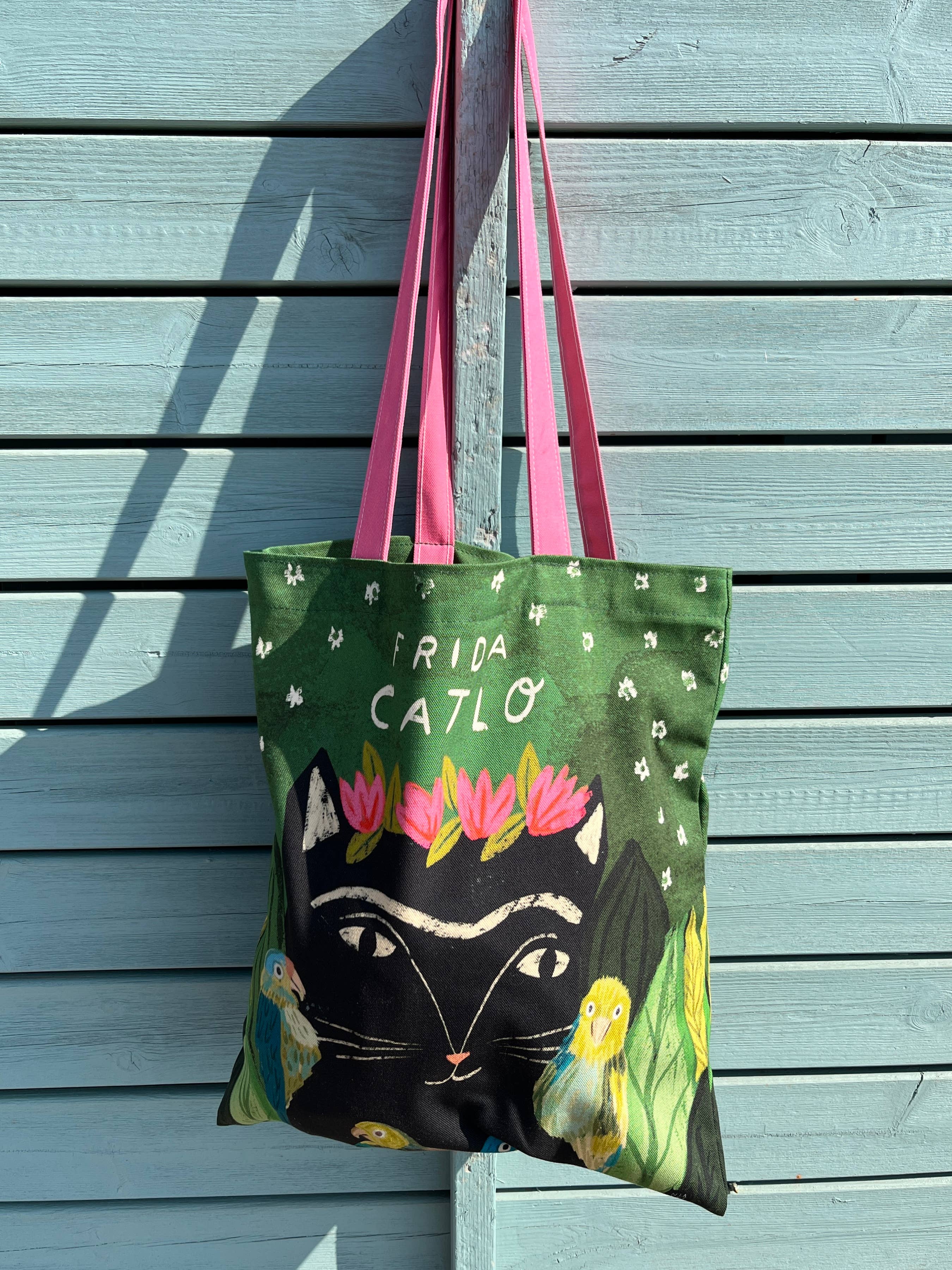 A tote bag with pink handles hanging against a light blue wooden wall. The bag features a green background with white text reading 'FRIDA CATLO', a large black cat face, pink and white flowers.
