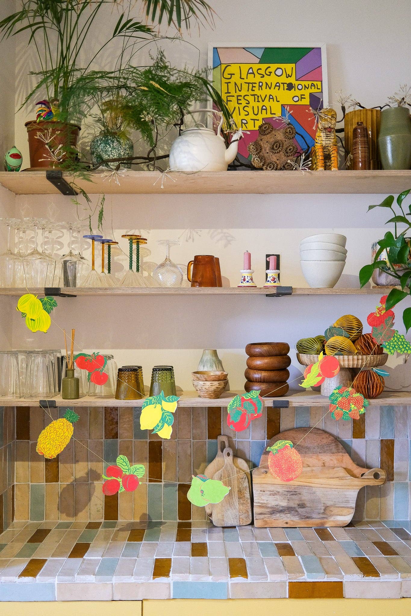 Shelves with decorative fruit garland, plants, and a colorful painting in a kitchen setting.