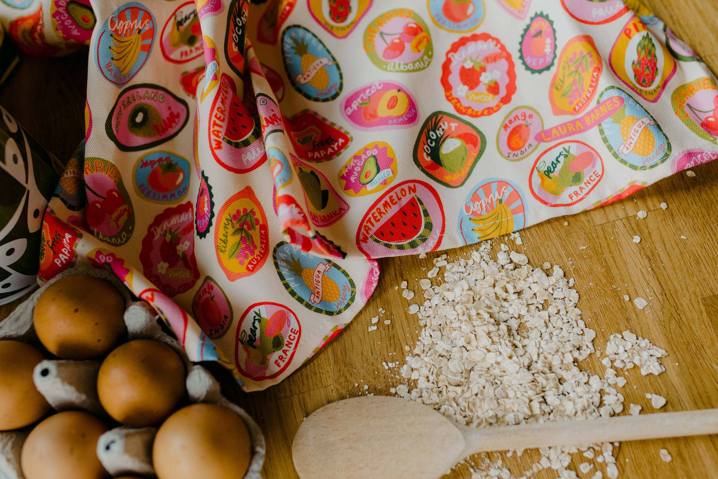 Colorful fabric with fruit patterns on a wooden surface with eggs and a wooden spoon.
