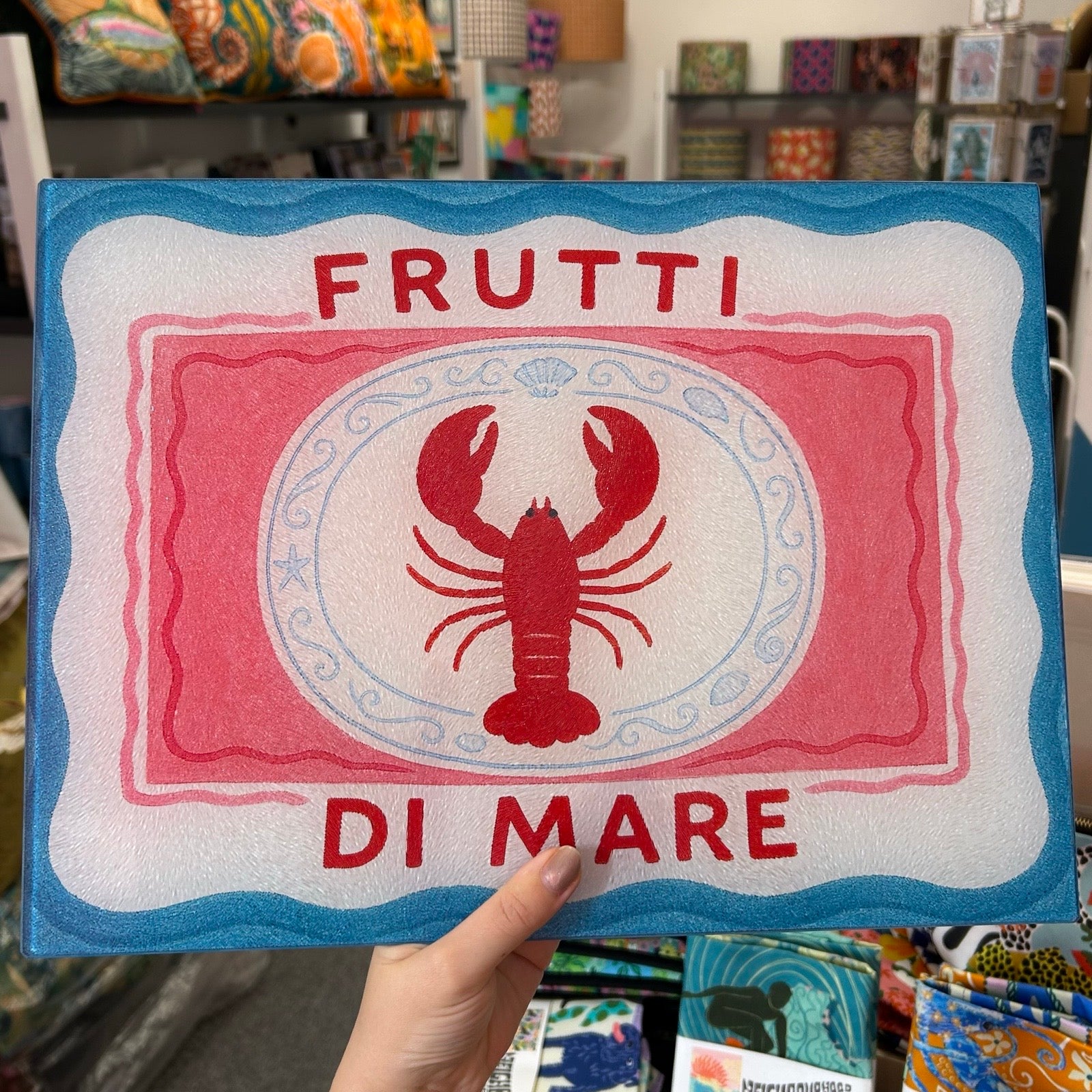 A hand holds a decorative chopping board featuring a red lobster in the centre, with the words “FRUTTI DI MARE” in red and a blue border, against a blurred store background.