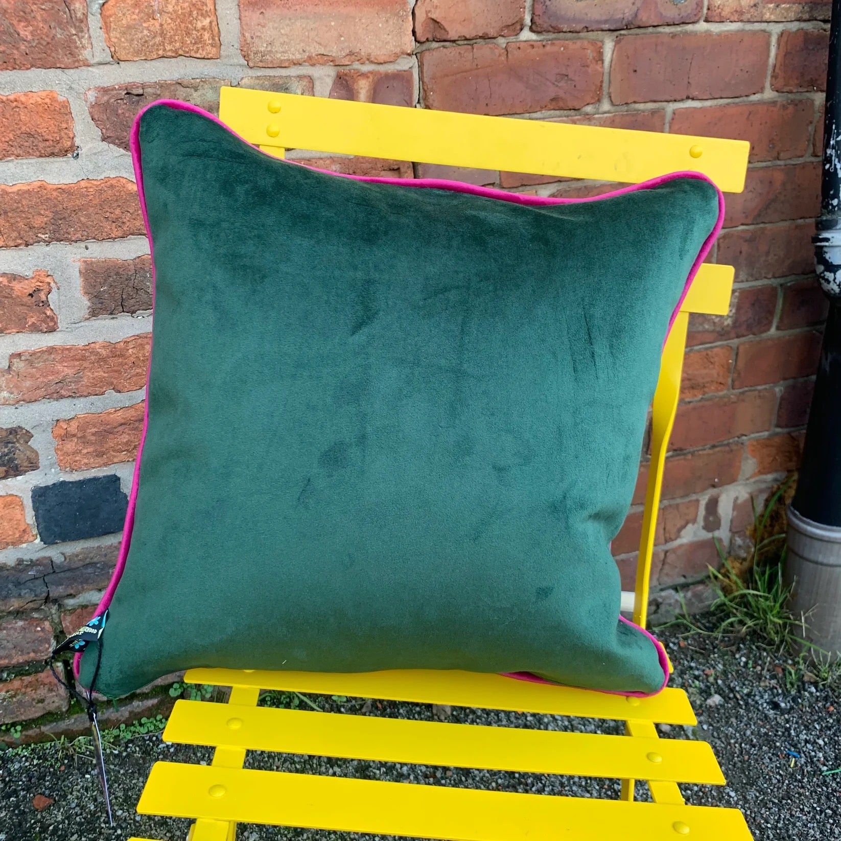 Green pillow with pink trim on a yellow chair against a brick wall.