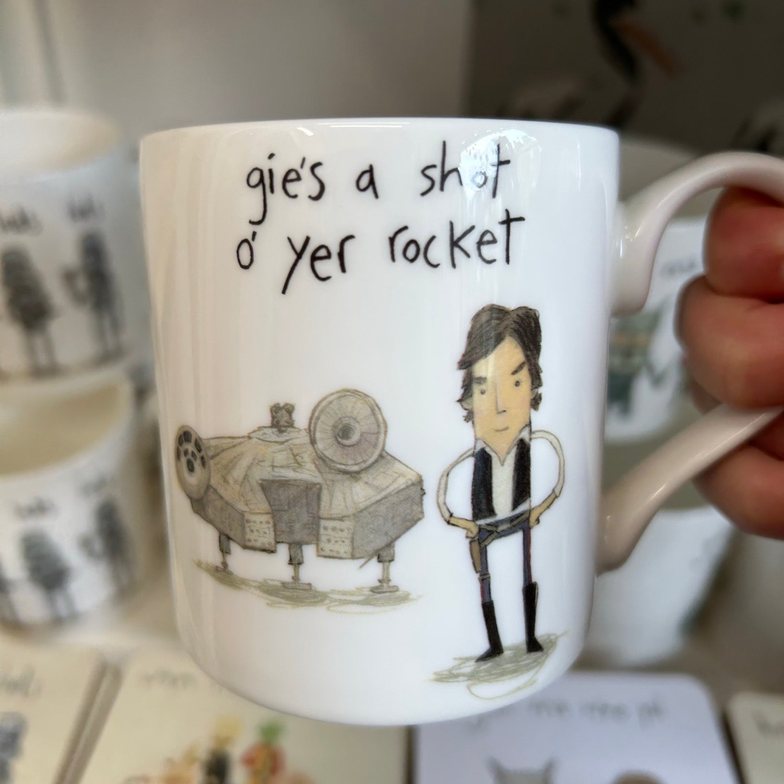 Mug with cartoon character and text 'gie's a shot o' yer rocket' held by a hand.