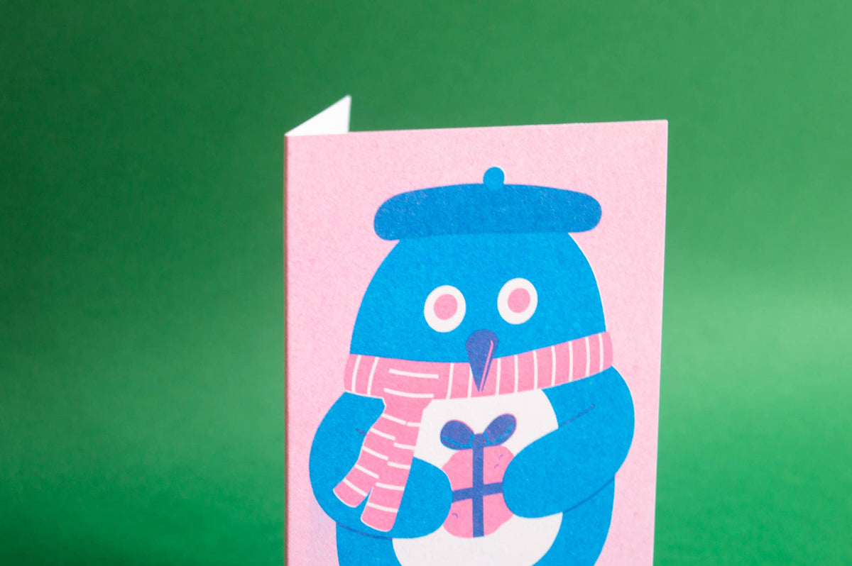 A pink greeting card featuring a blue penguin wearing a pink striped scarf and holding a gift, photographed against a solid green background.
