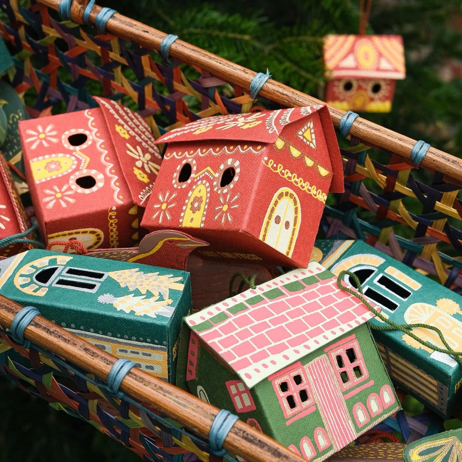 Colourful paper house ornaments in a basket with a Christmas tree in the background.