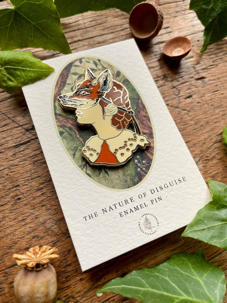 Enamel pin of a fox and face on a card with green leaves and wooden background.