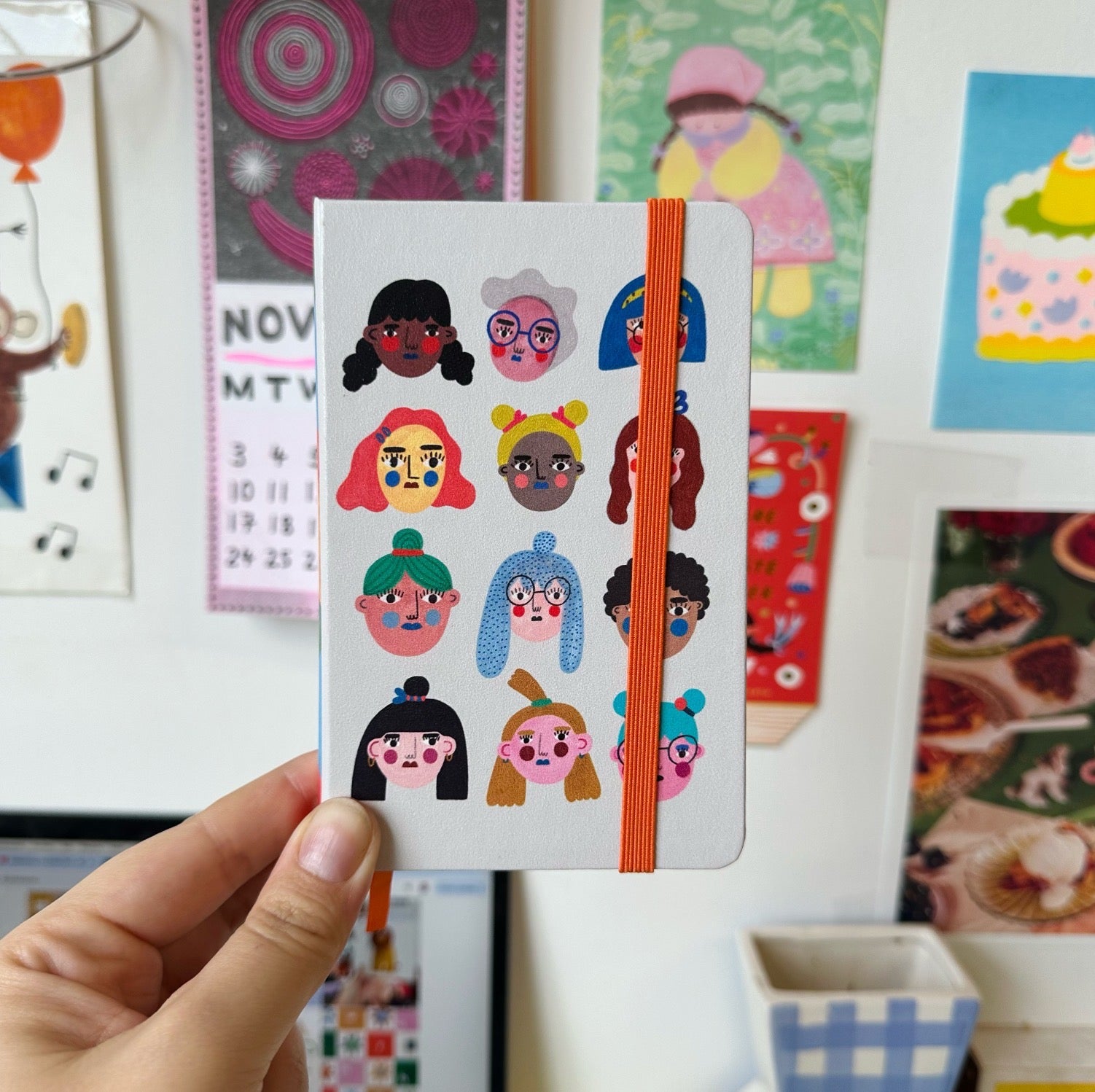 Notebook with colourful character design held in front of a wall with various posters and decorations.