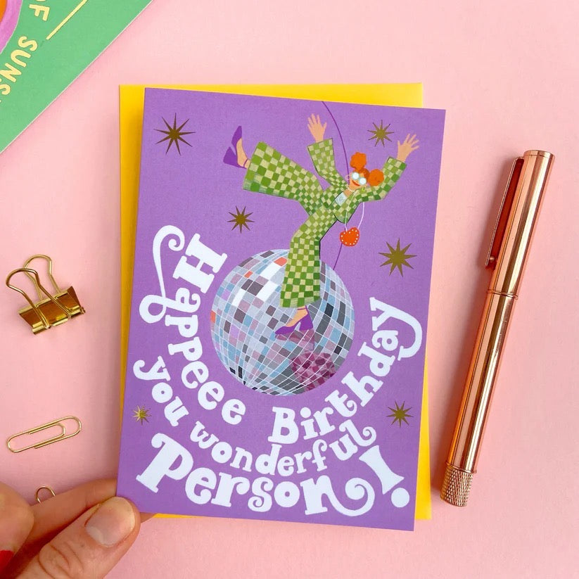 Birthday card with disco ball and a woman in a green jumpsuit on a pink background.