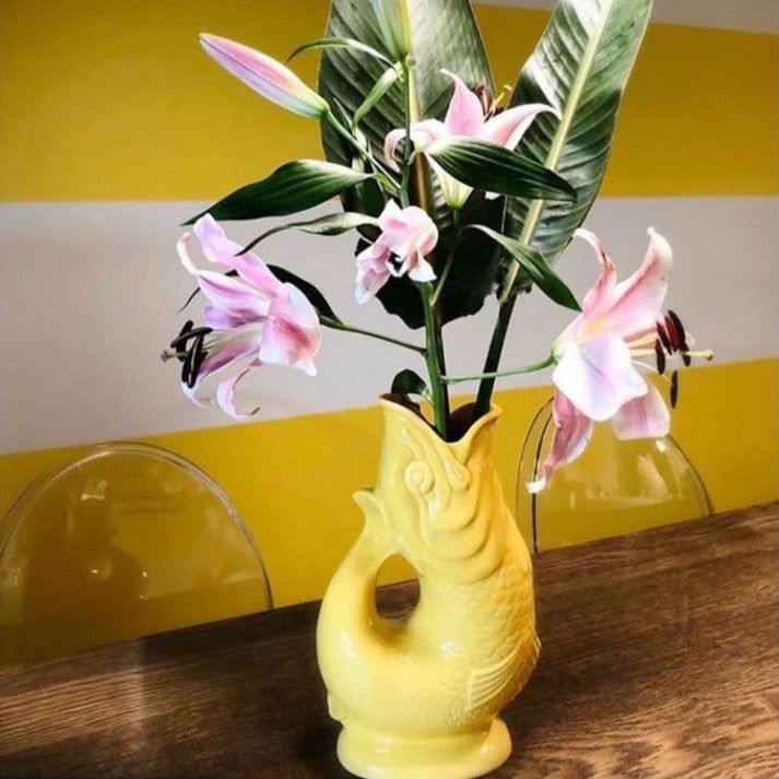 Yellow fish-shaped jug with pink flowers on a wooden surface against a yellow and white striped wall.