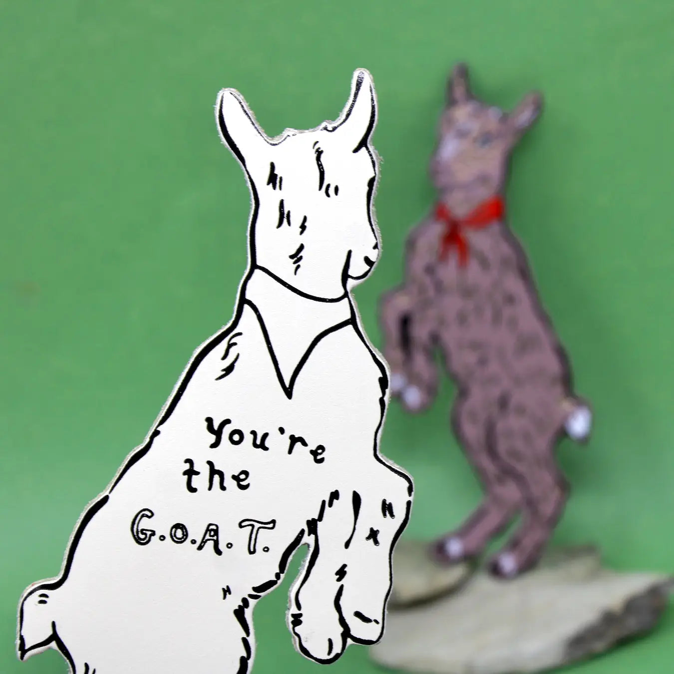 Bookmark of a goat with 'You're the G.O.A.T.' text next to a small figurine of a goat on a green background.