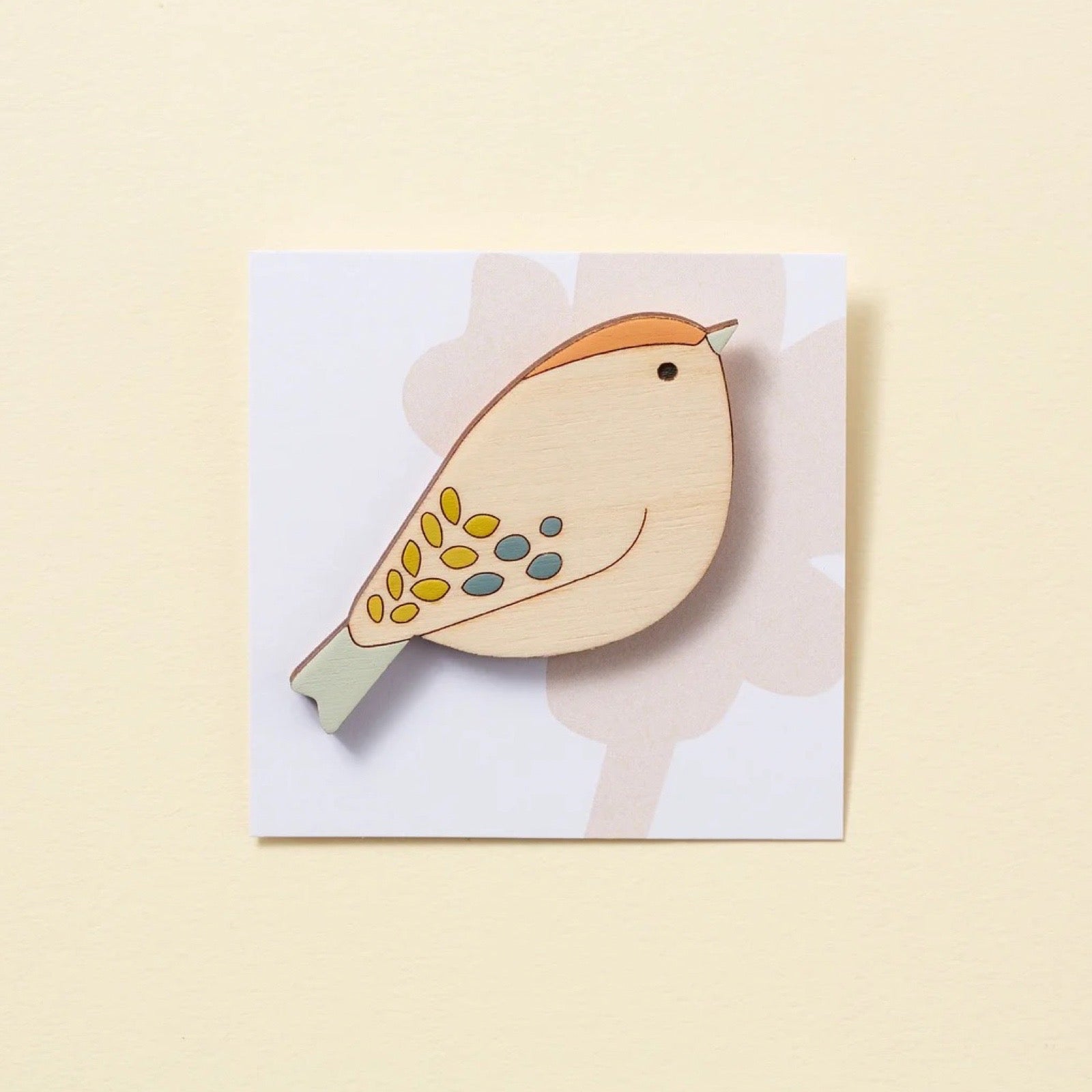 Wooden bird brooch on a light pink card on a yellow background.
