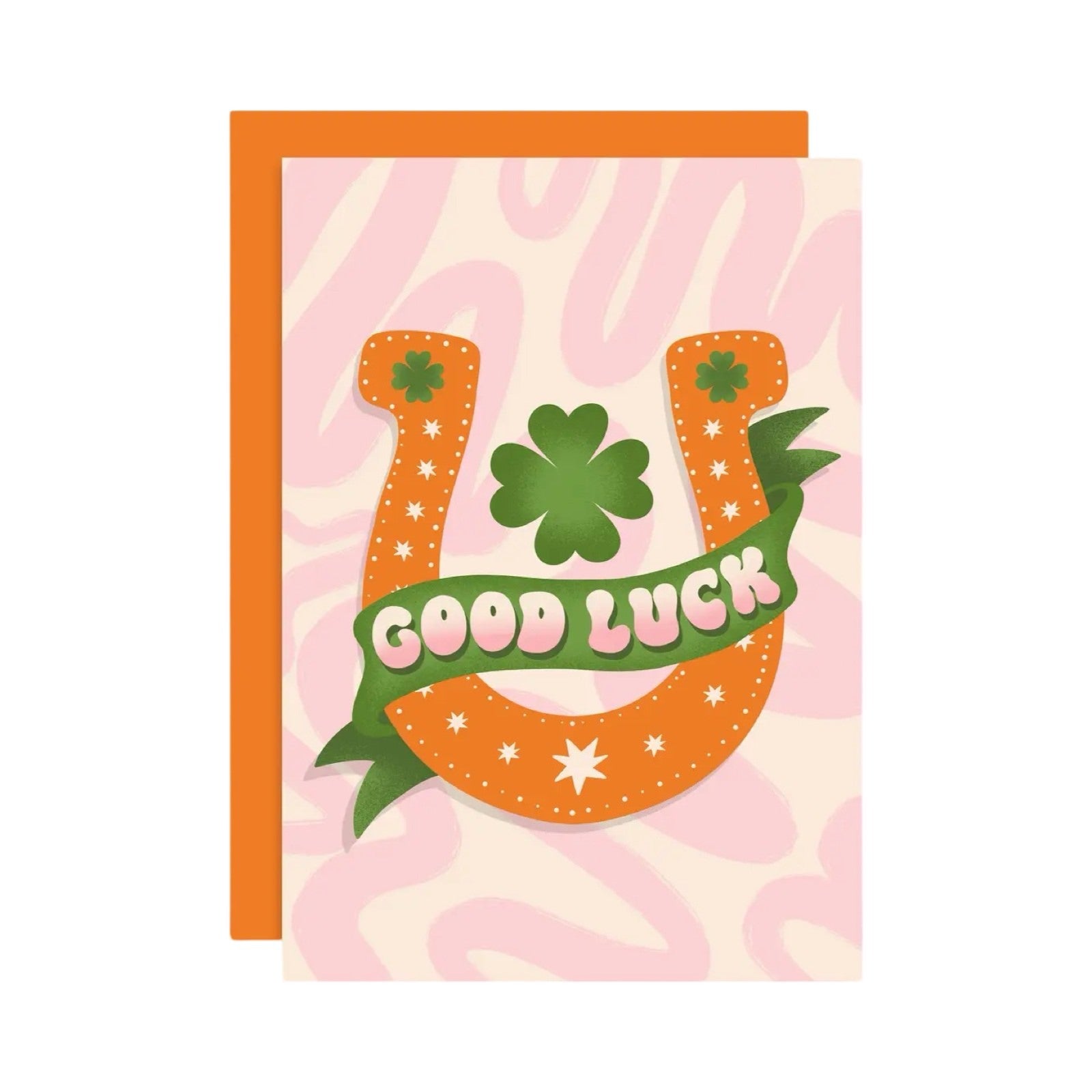 A greeting card with an orange horseshoe and a green four-leaf clover on a pink and cream background, with the text 'Good Luck' in a green banner.