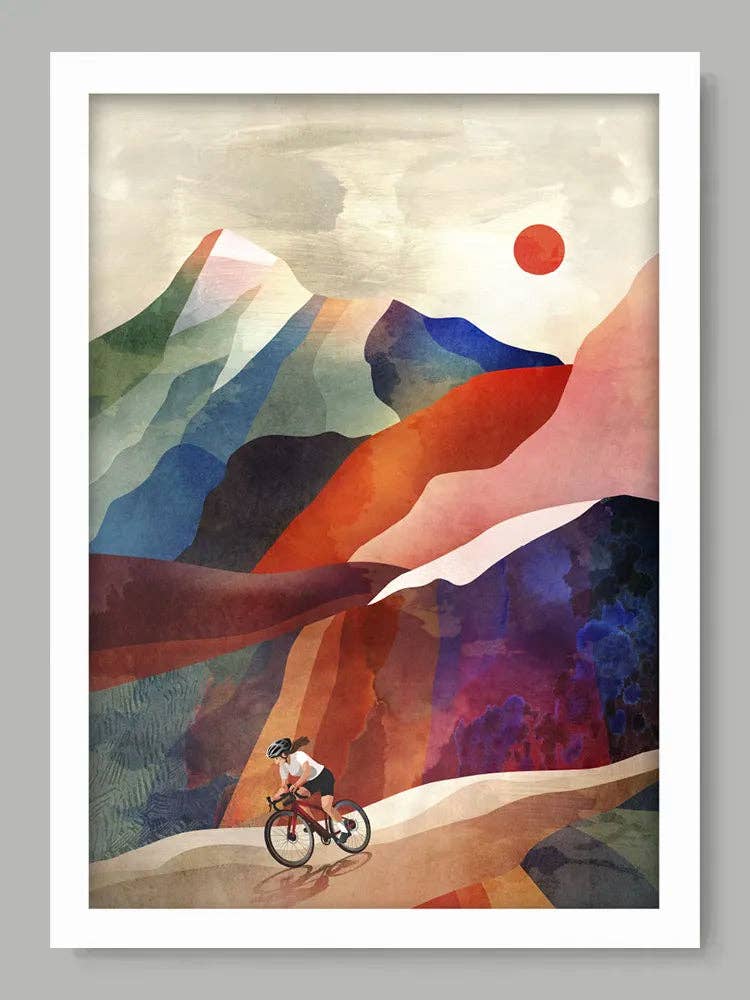 A framed art print featuring an abstract, colorful landscape with layered mountains in shades of blue, purple, orange, and green, a small cyclist near the bottom, and a red sun in the sky. The print is presented in a white frame against a neutral grey background.