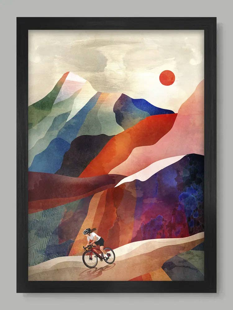 Framed abstract landscape art print featuring stylized mountains, a cyclist on a winding path, and a red sun, shown in a black frame against a light grey background.