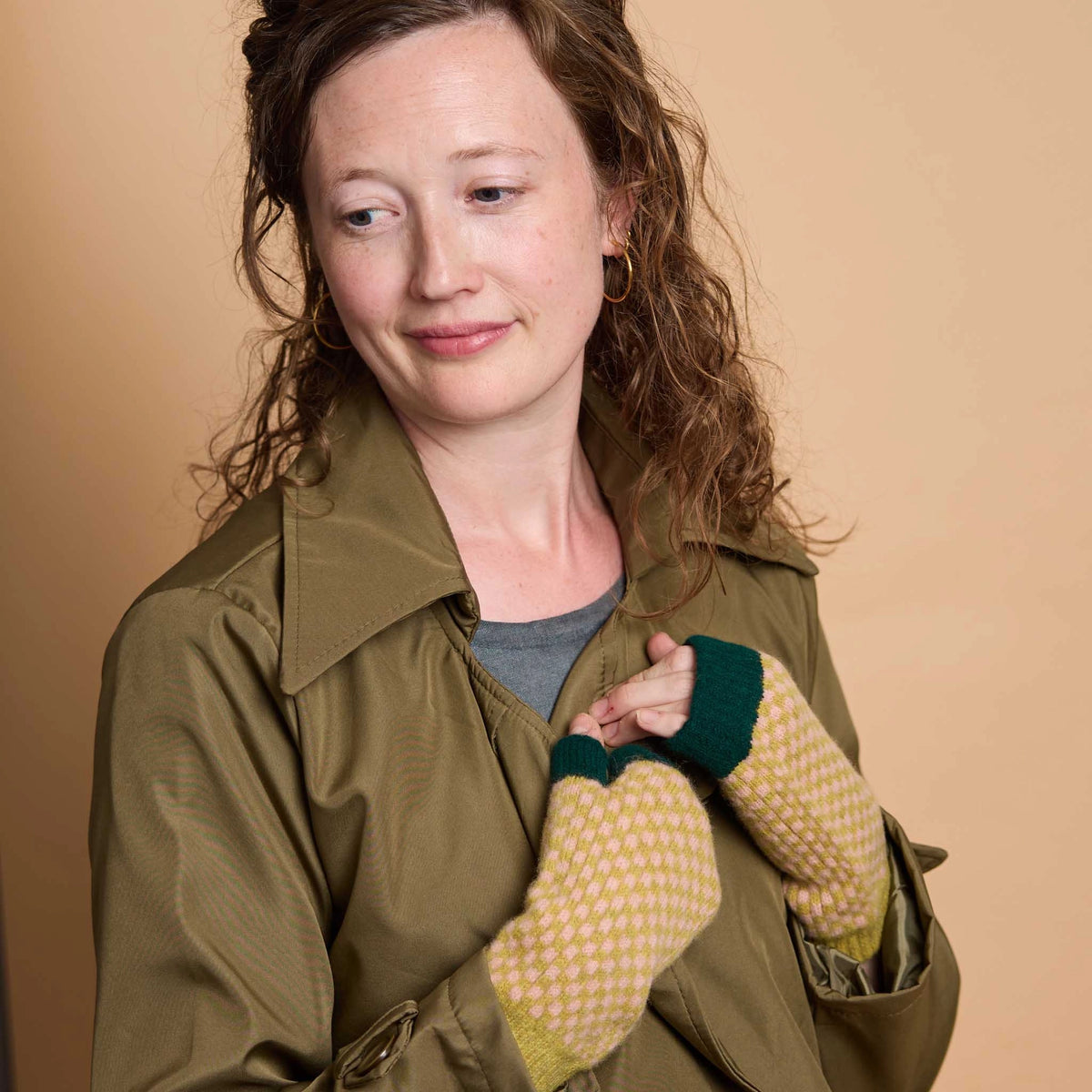 A woman wearing a green jacket and yellow-and-white checkered fingerless gloves with dark green cuffs, posed against a solid beige background.