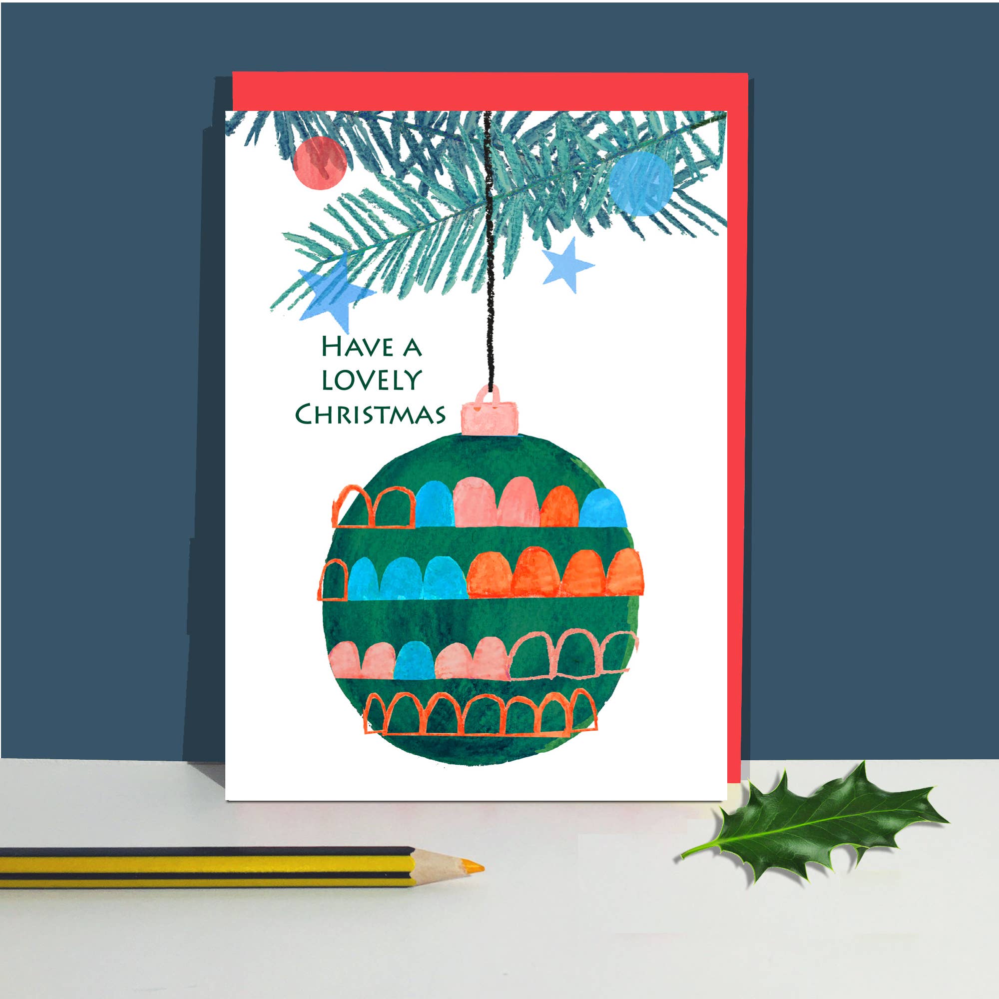 A Christmas greeting card with a white background, featuring a colorful watercolor ornament illustration, pine branches, stars, and the text 'HAVE A LOVELY CHRISTMAS'. The card is shown standing upright on a white surface against a dark blue background, with a pencil and holly leaf nearby.