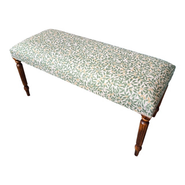 A rectangular bench with mahogany legs and cream linen fabric upholstered in a green foliage pattern.