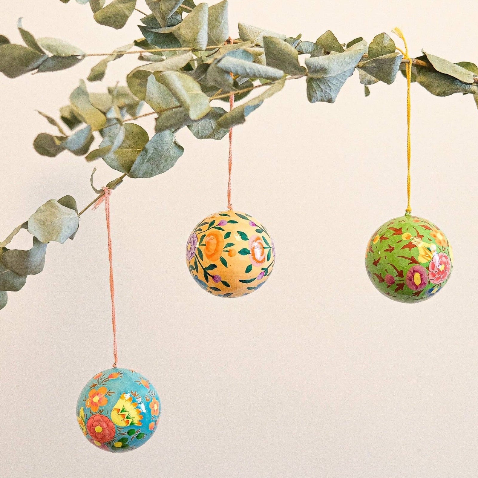 3 round papier mache baubles hanging on a branch, one green floral bauble, one turquoise floral bauble and one peach floral bauble.