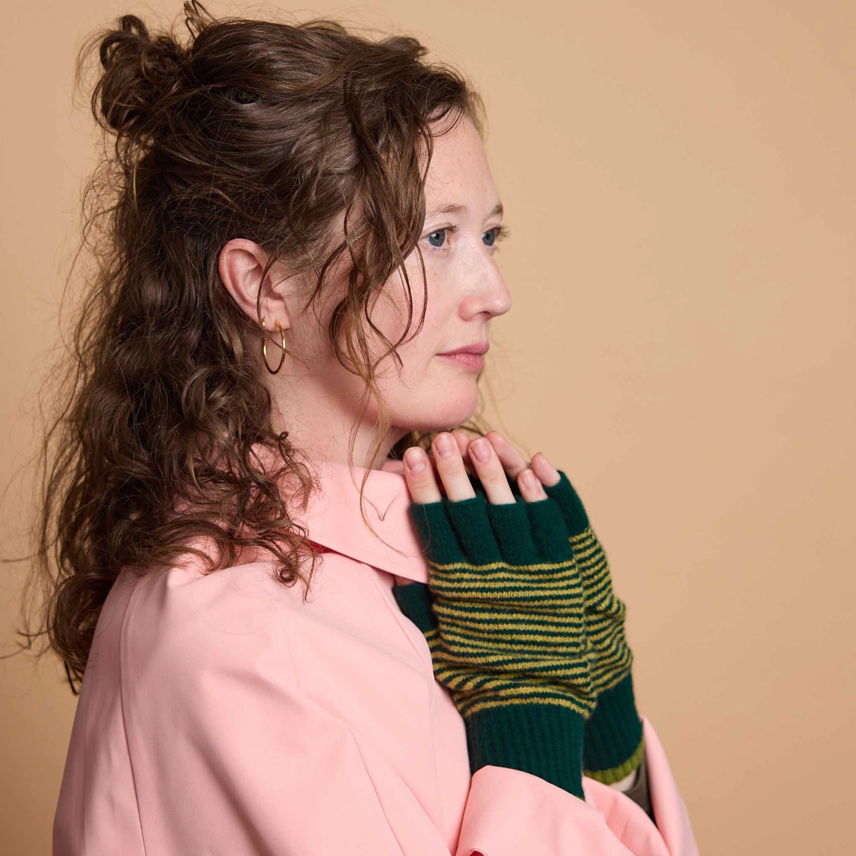 A woman in a pink shirt wearing dark green knit fingerless gloves with yellow stripes, posed against a solid beige background.