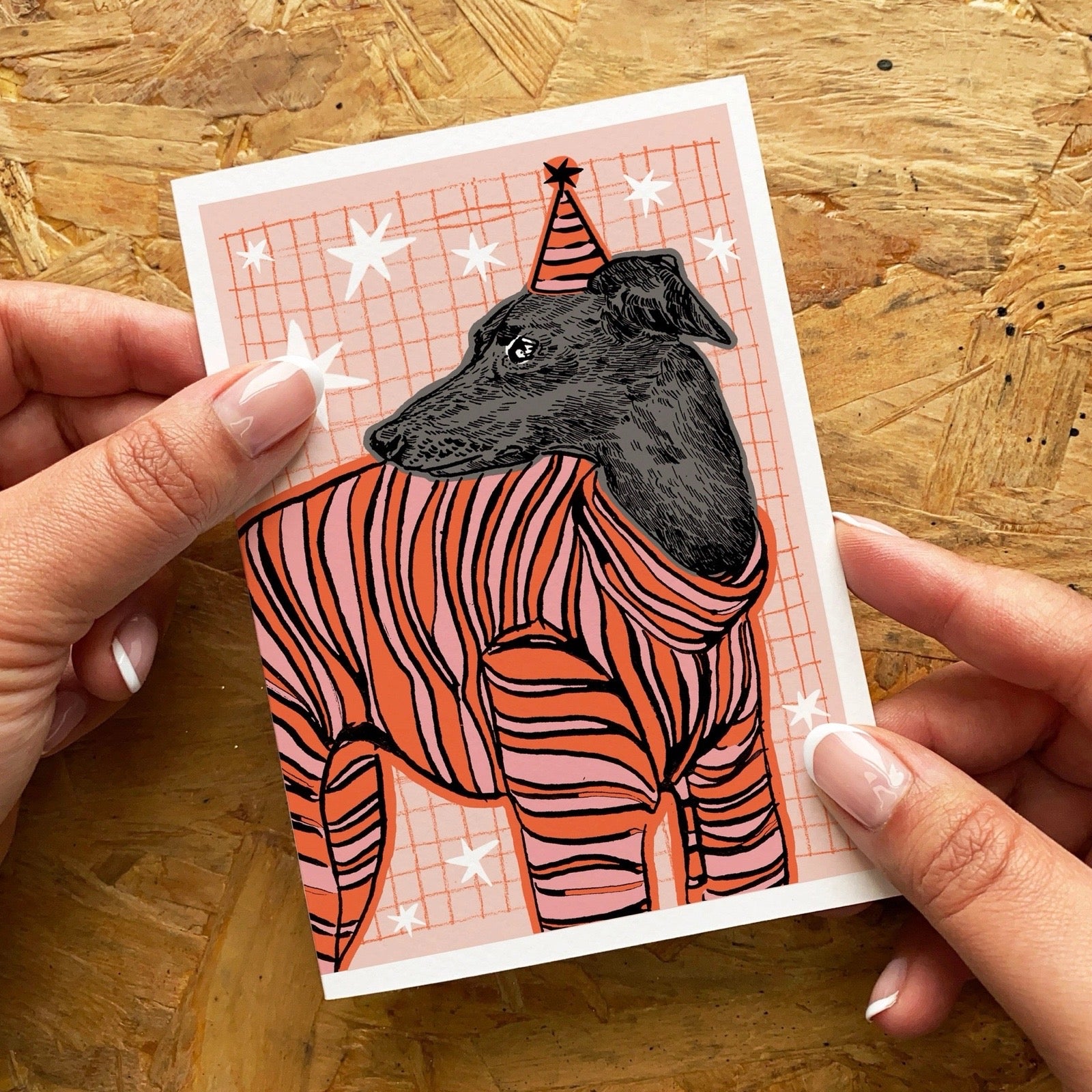 Illustration of a dog in a stripe jumper on a card held by hands against a wooden background.