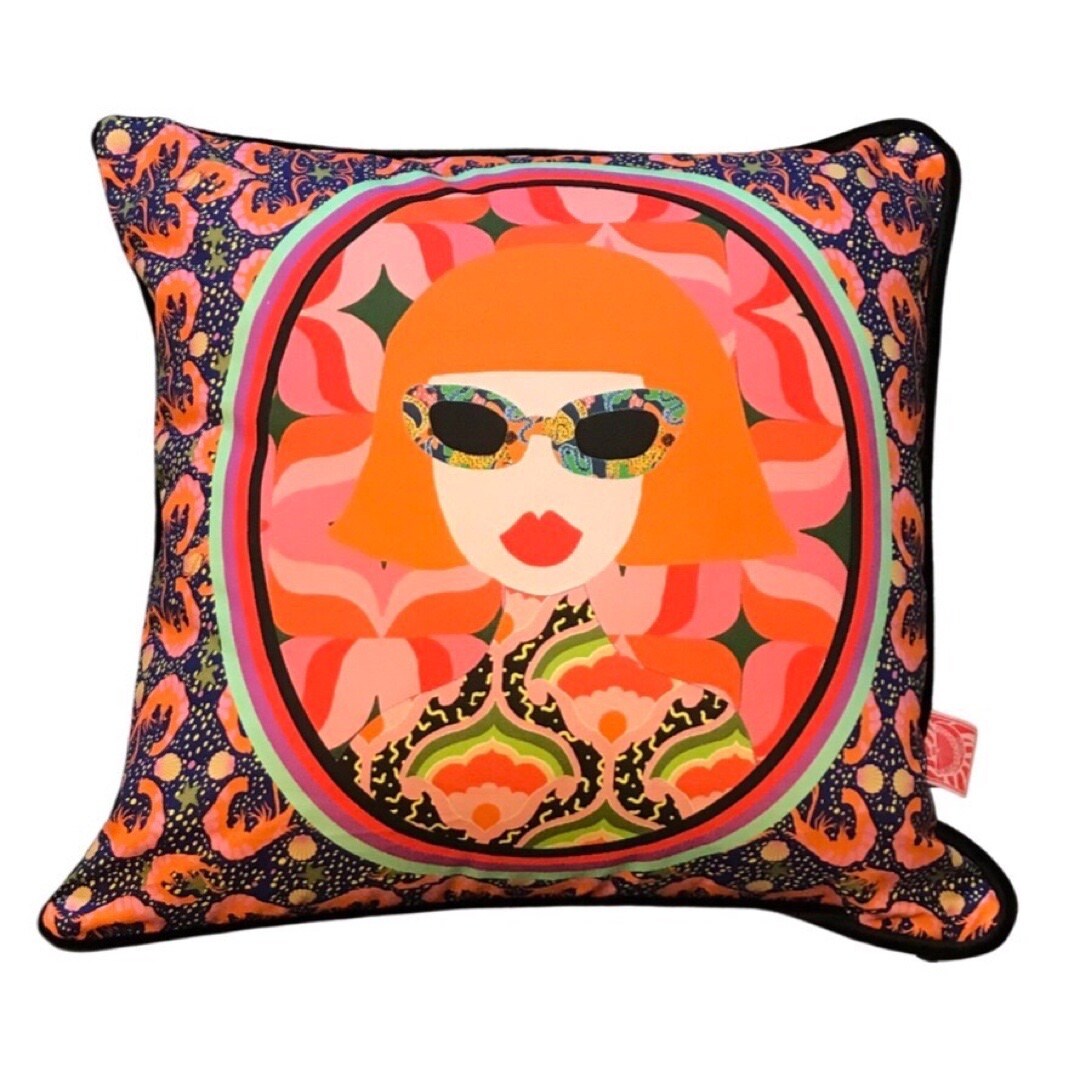 A cushion with an illustration of a seventies-inspired lady's face with gorgeous red lips, orange hair and multicoloured sunglasses circled within a pink and red patterned circle with an illustrated shrimp background.